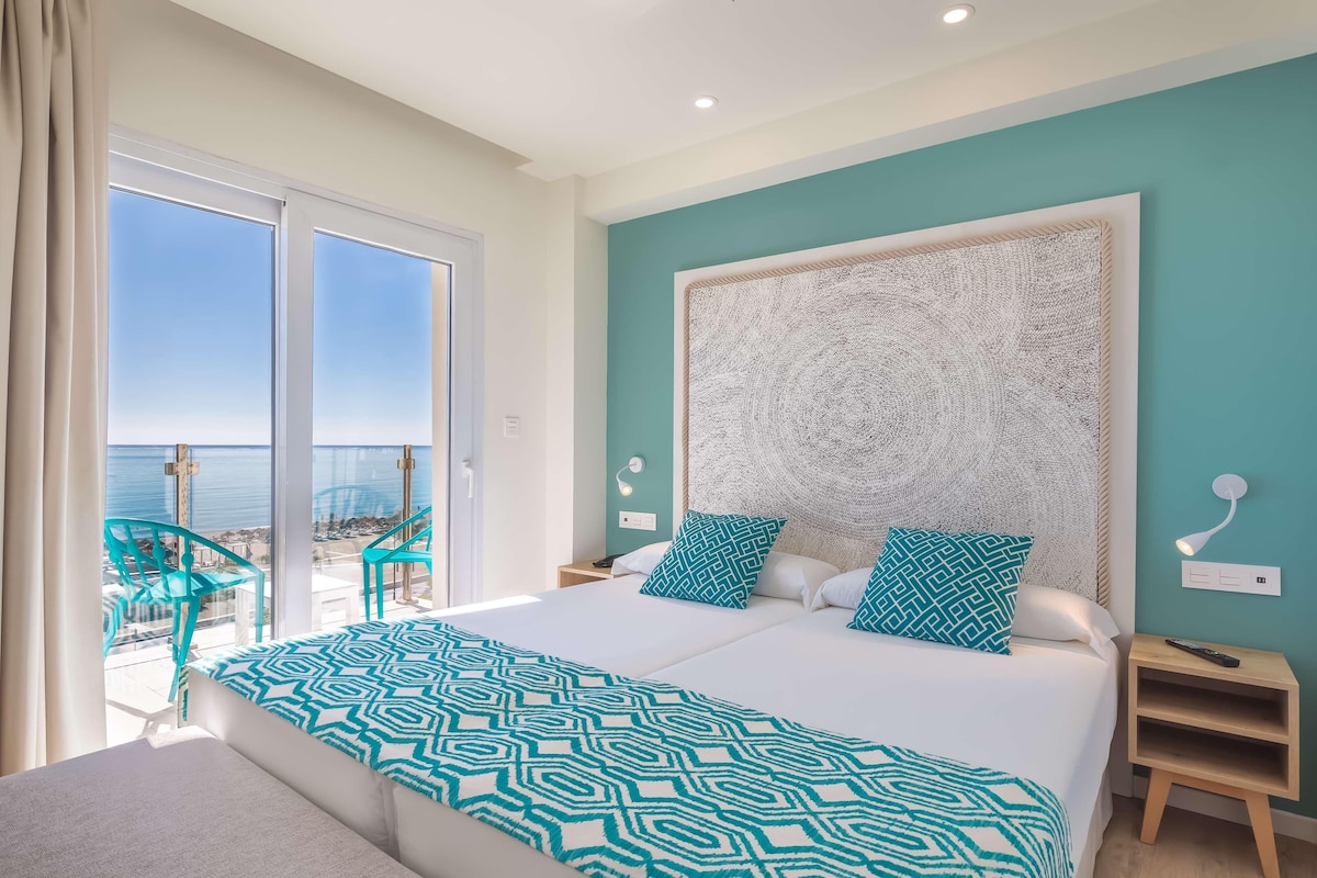 A bedroom with a large bed, two nightstands, a view of the sea, and a balcony with chairs.