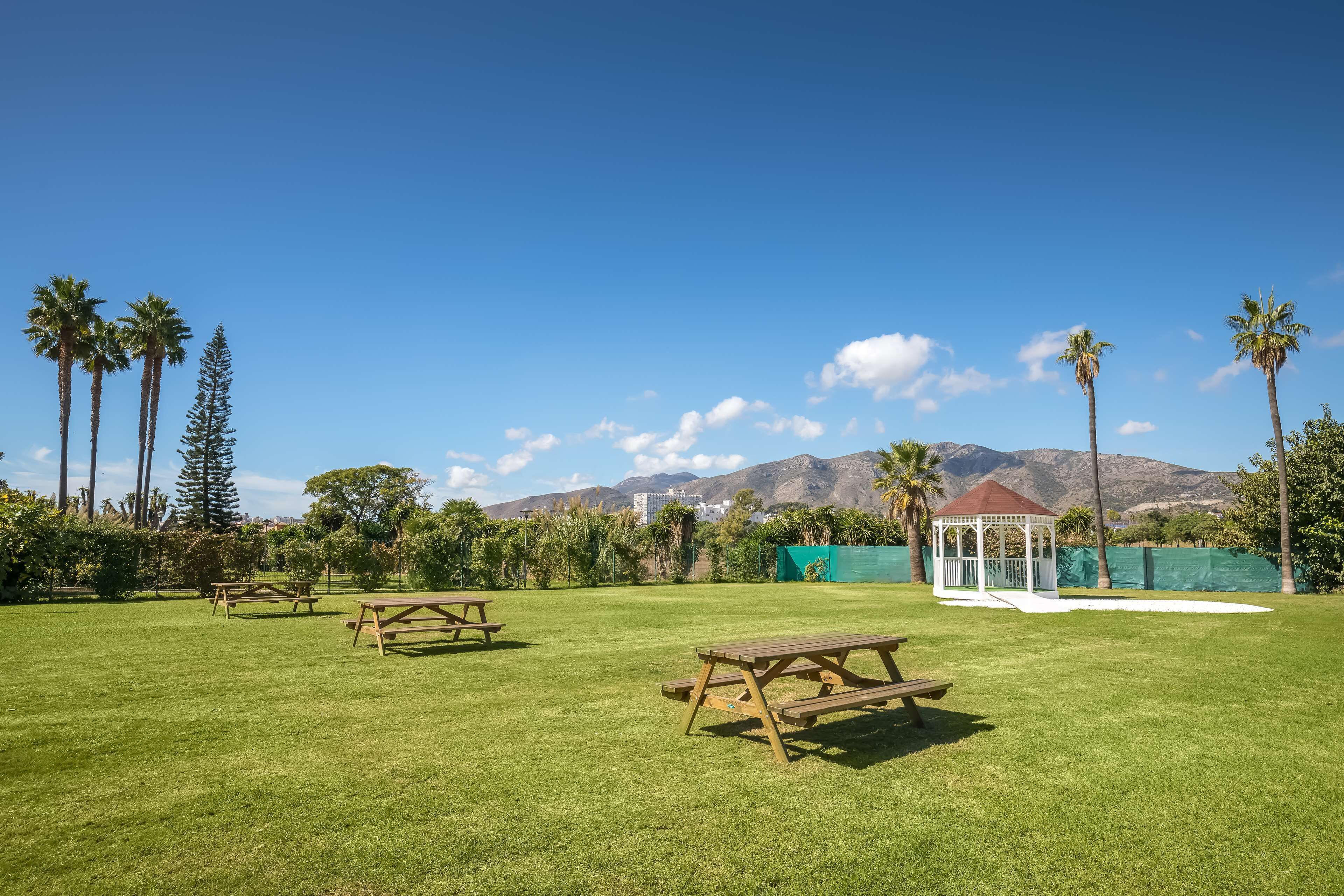 Un'area verde con tavoli da picnic e un gazebo, circondata da palme e montagne sullo sfondo.