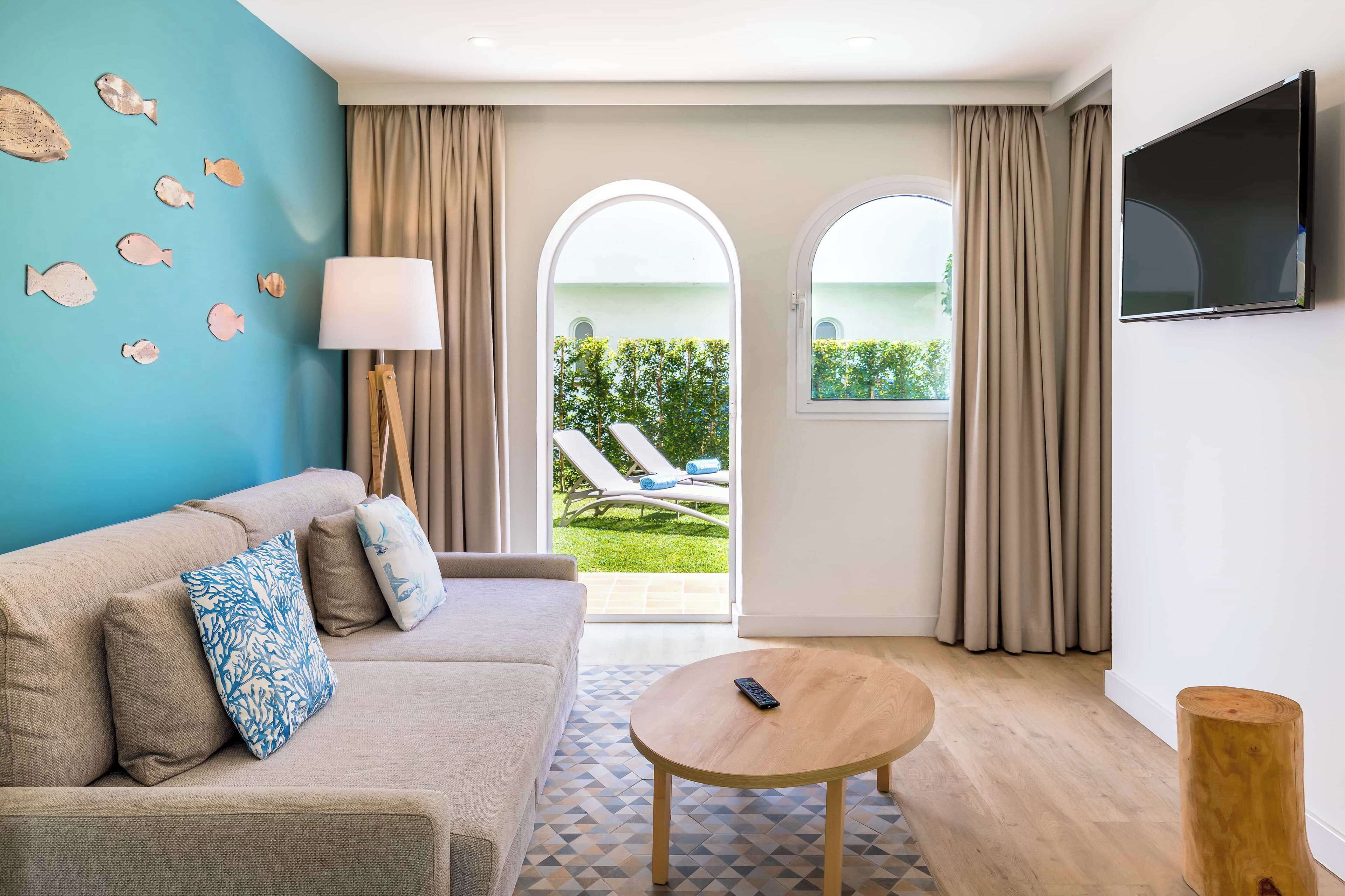 Ein modernes Hotelzimmer mit einem beigen Sofa, einem runden Holztisch, einem an der Wand montierten Fernseher und Blick auf einen Garten mit Pool und Liegestühlen.
