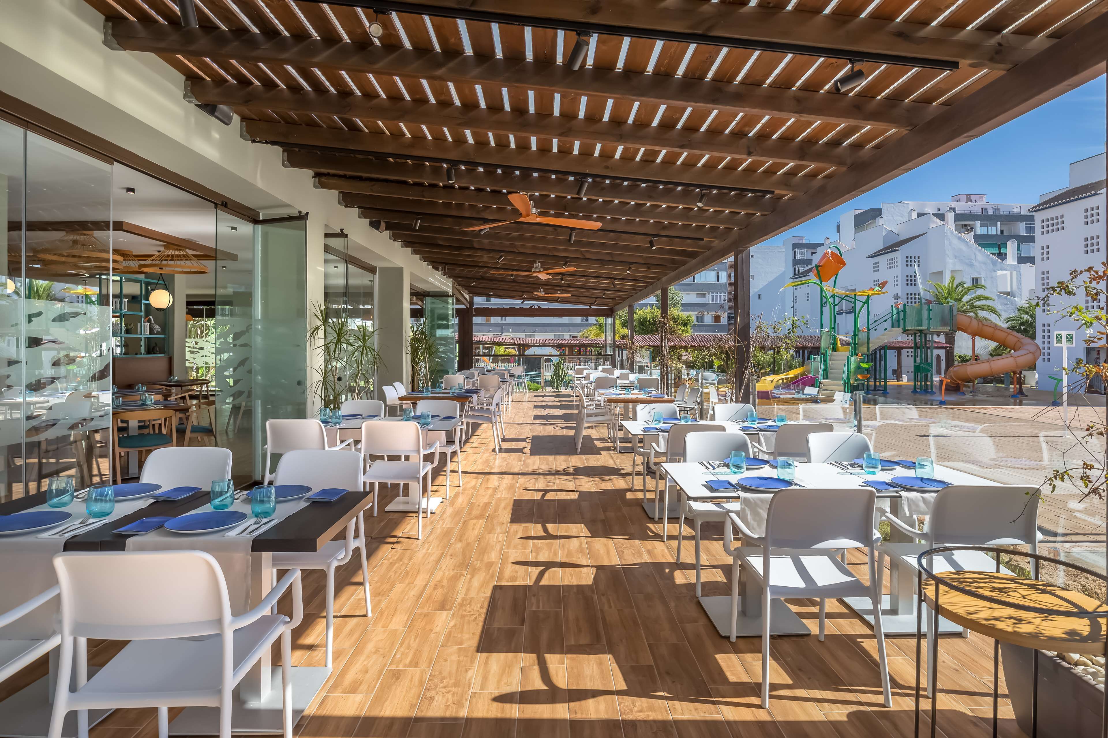 Un área de comedor al aire libre con piso de madera, sillas blancas y mesas dispuestas para comer.