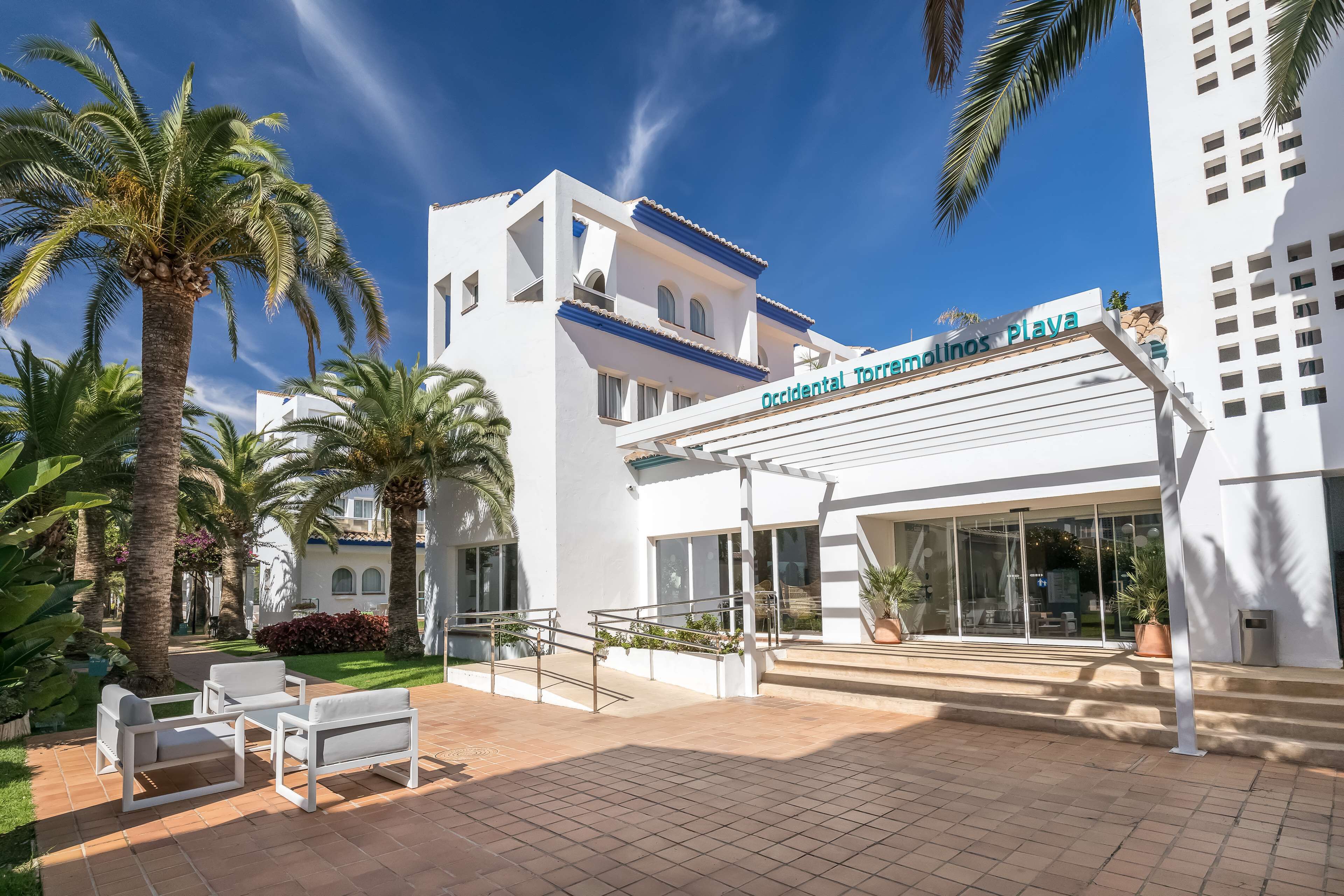 A modern hotel with a white facade, palm trees, and a clear blue sky.