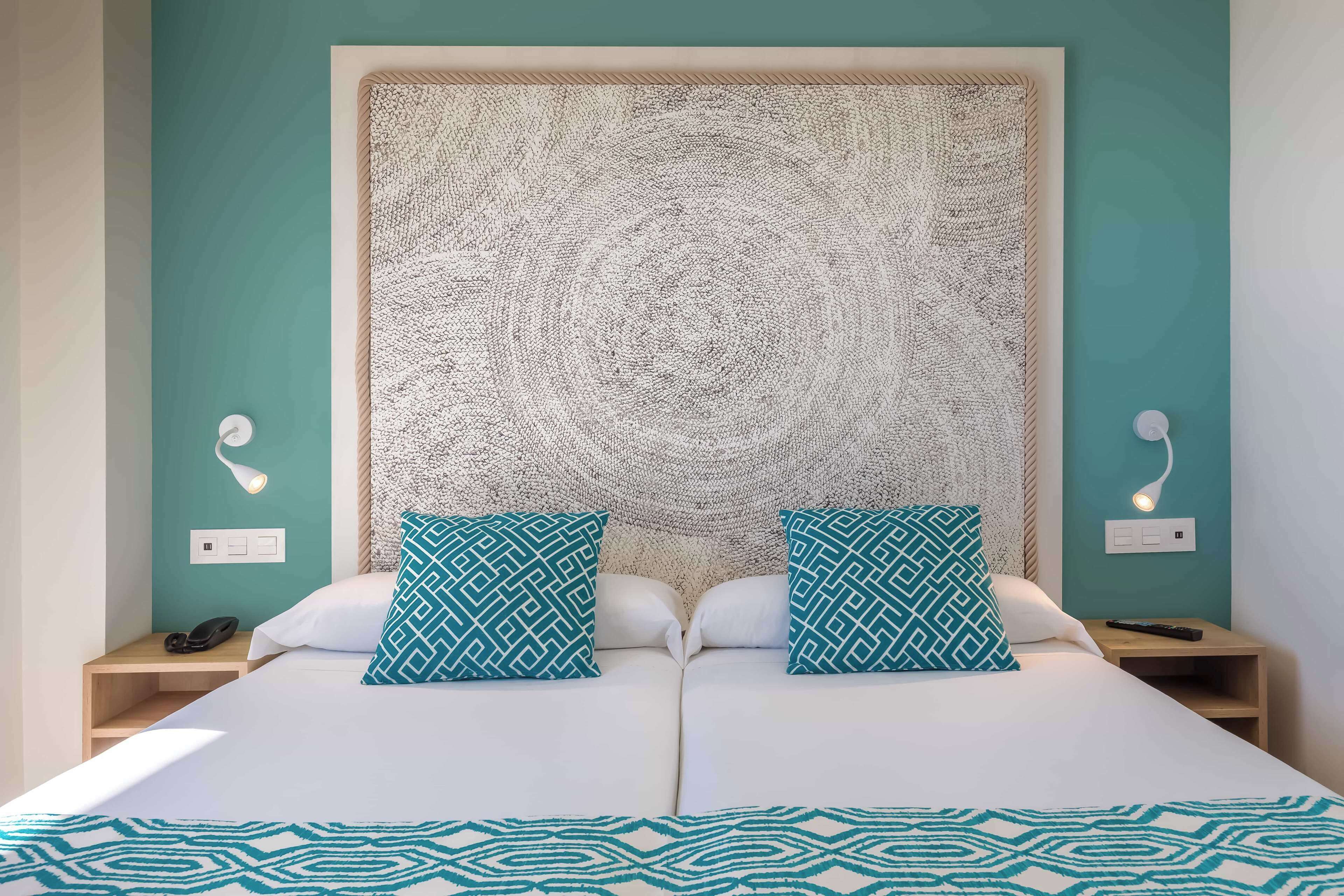 A double bed with a patterned bedspread, two matching pillows, a textured headboard, bedside lamps, and a nightstand with a phone.