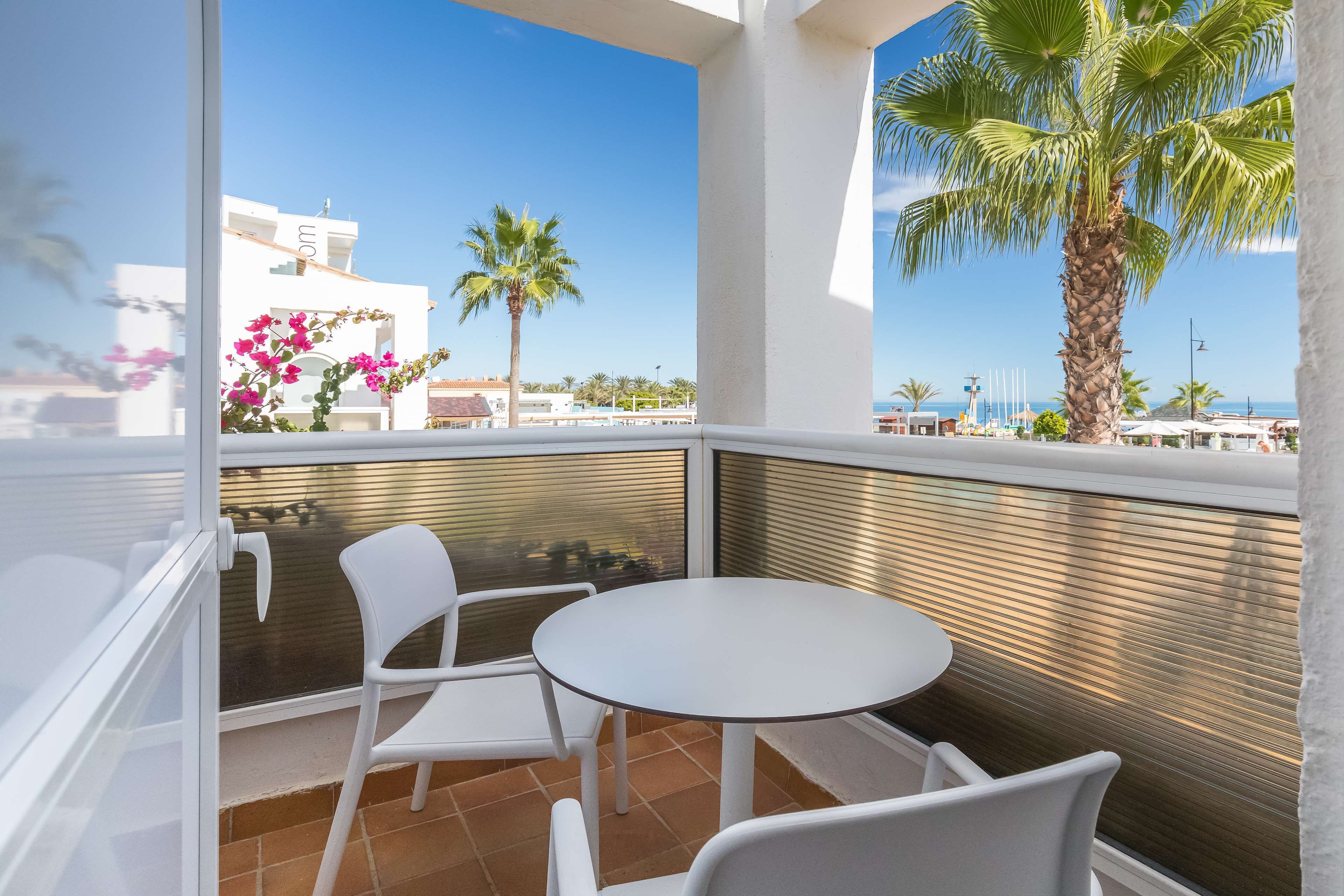 Un balcon avec des chaises blanches et une table, donnant sur une plage bordée de palmiers et surplombant un ciel dégagé.