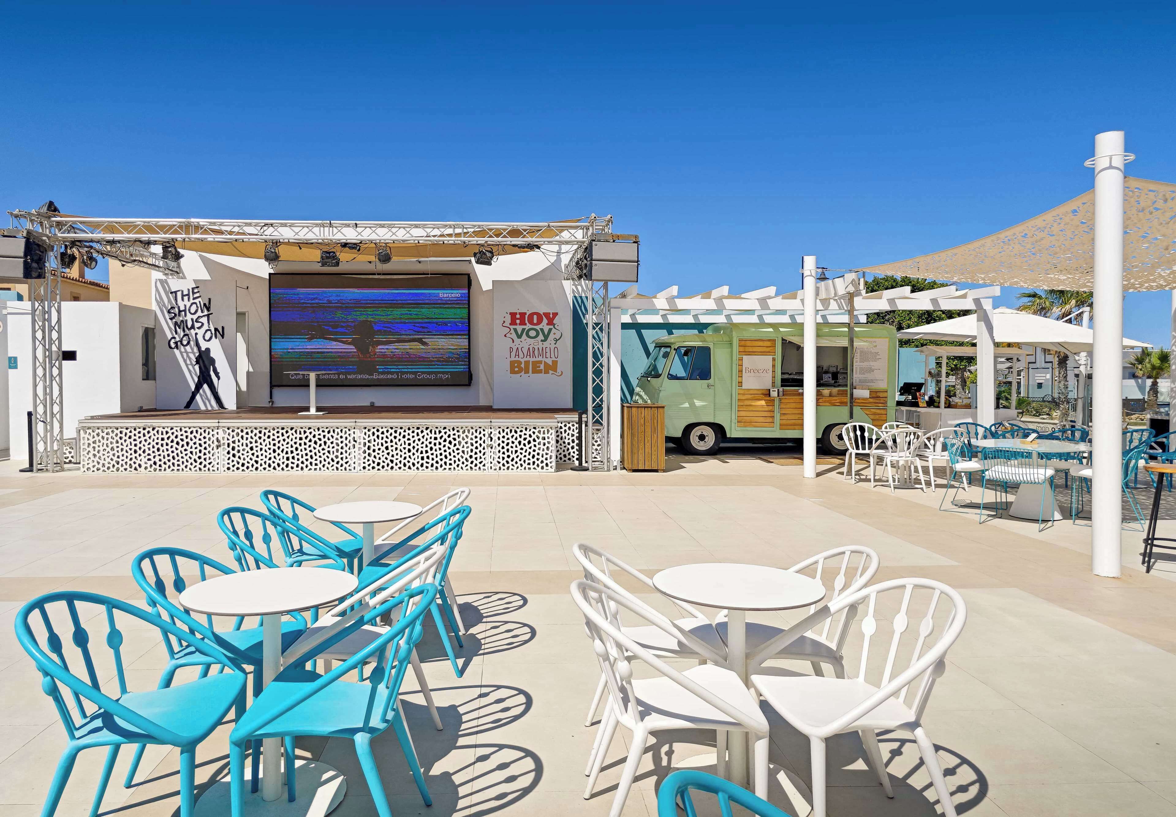 An outdoor seating area with tables and chairs, a stage with a screen, and a food truck.