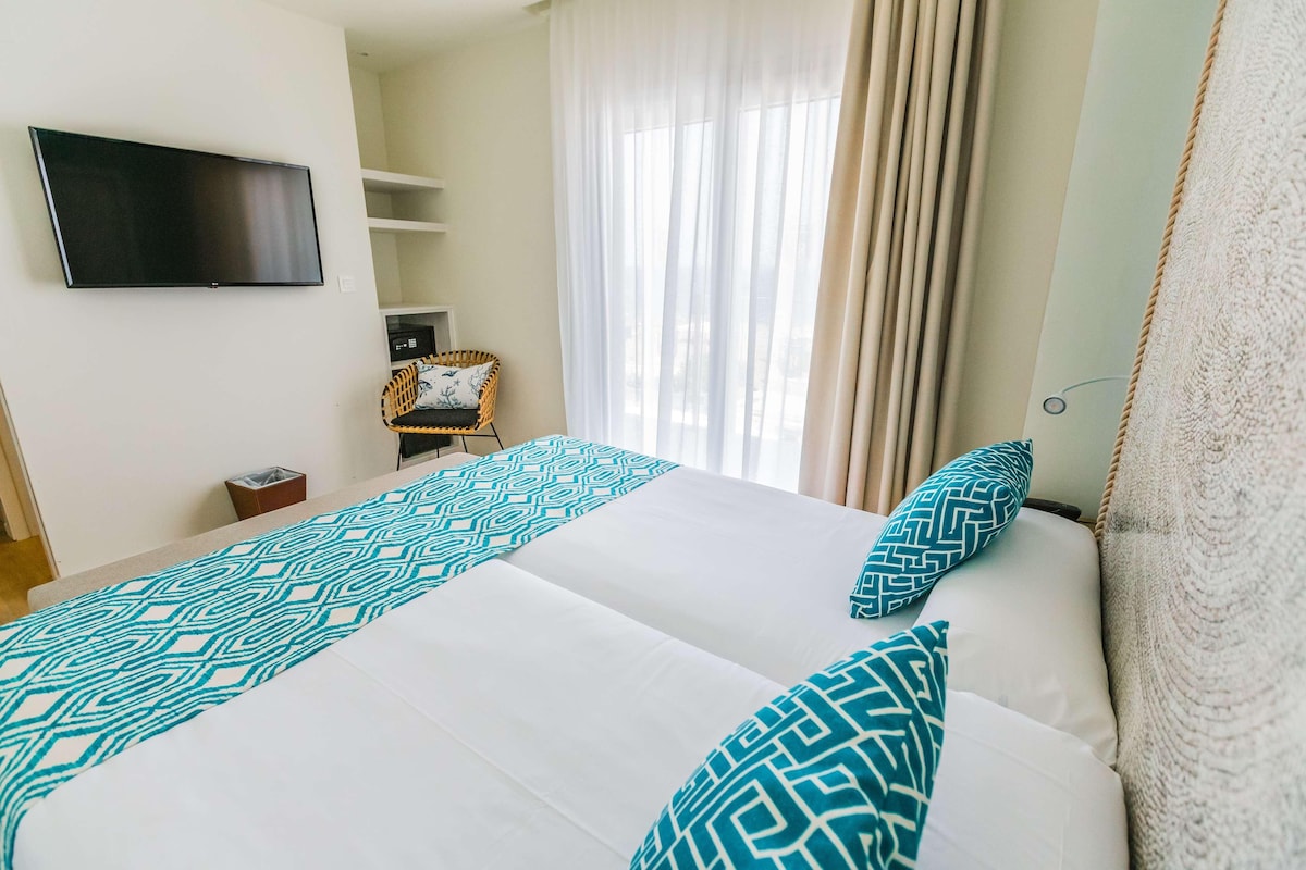 A hotel room with two beds, a TV mounted on the wall, a window with curtains, and a small table with a basket.