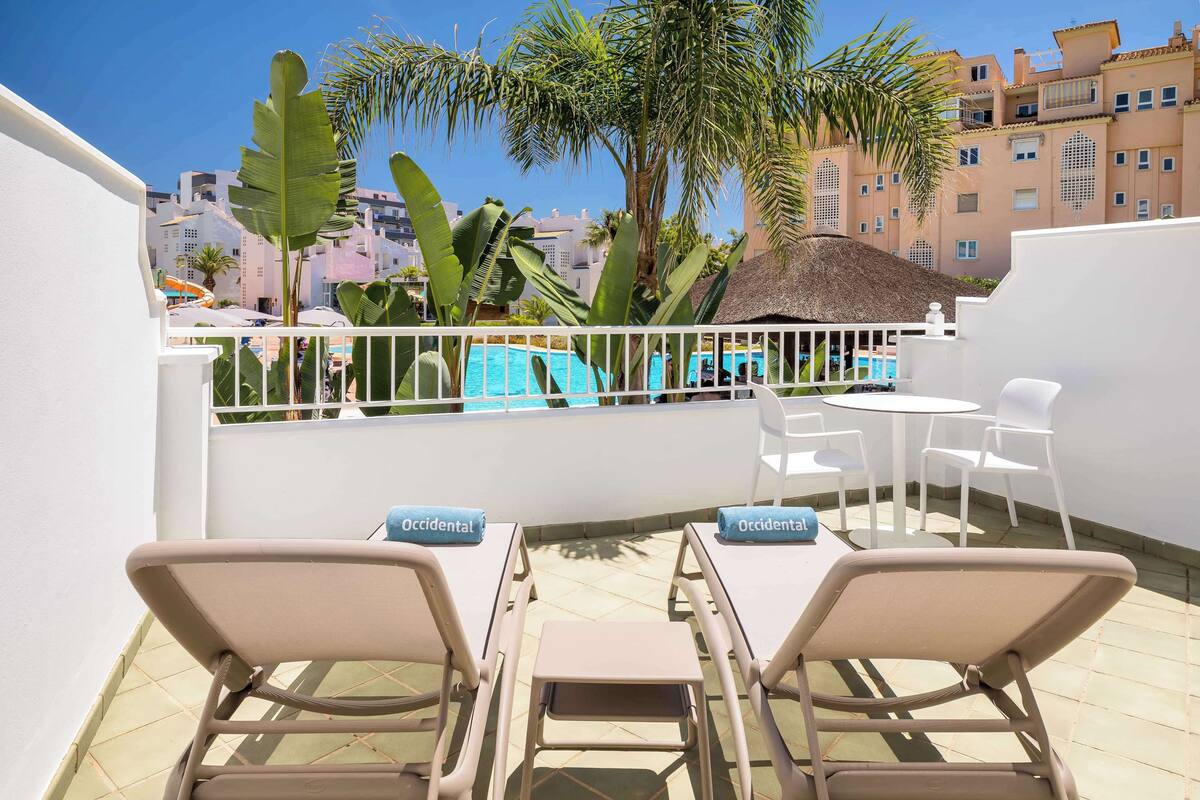 A rooftop terrace with two lounge chairs, a small table, and a view of a swimming pool.