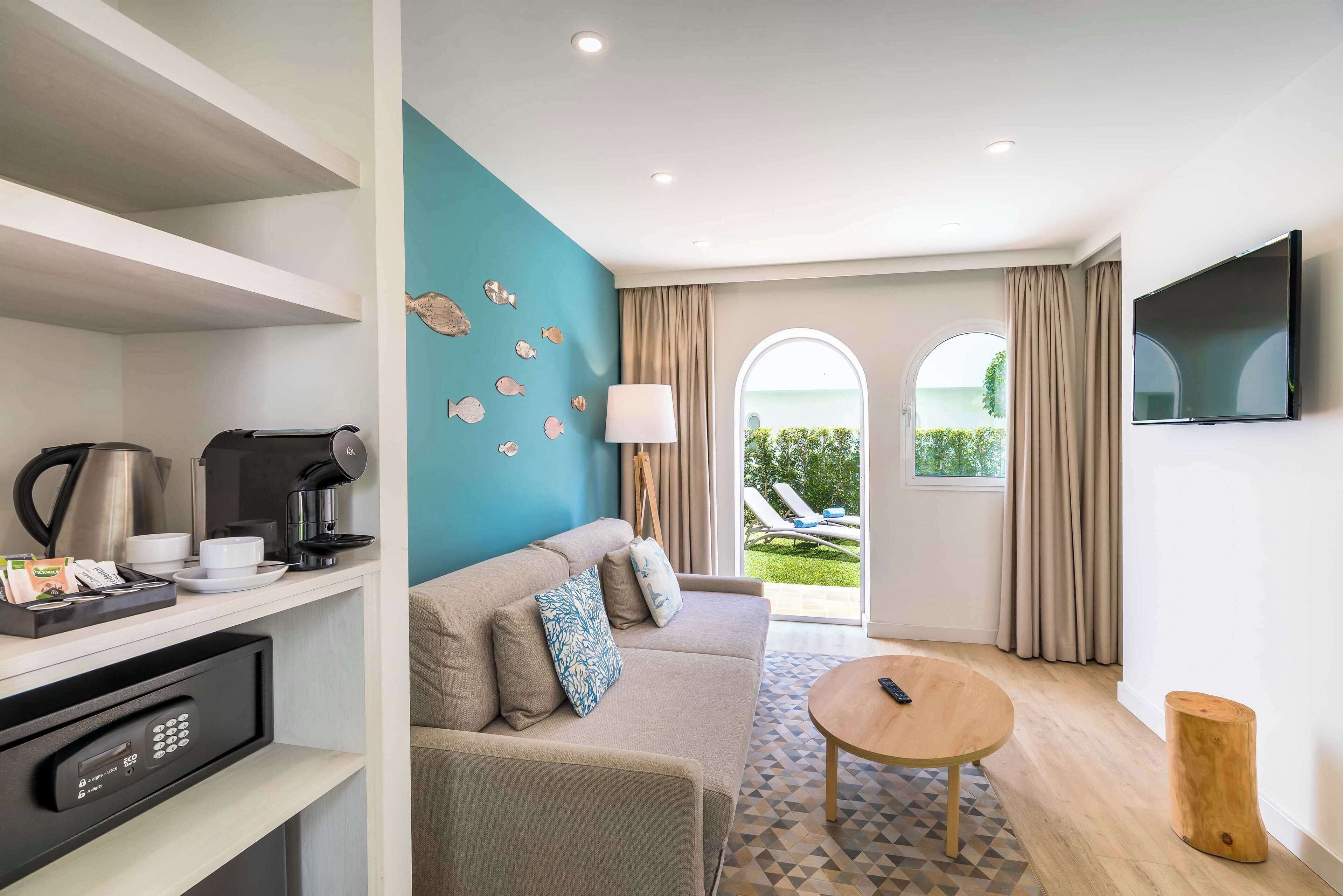 A modern hotel room with a sofa, coffee station, TV, and a view of the outdoors.