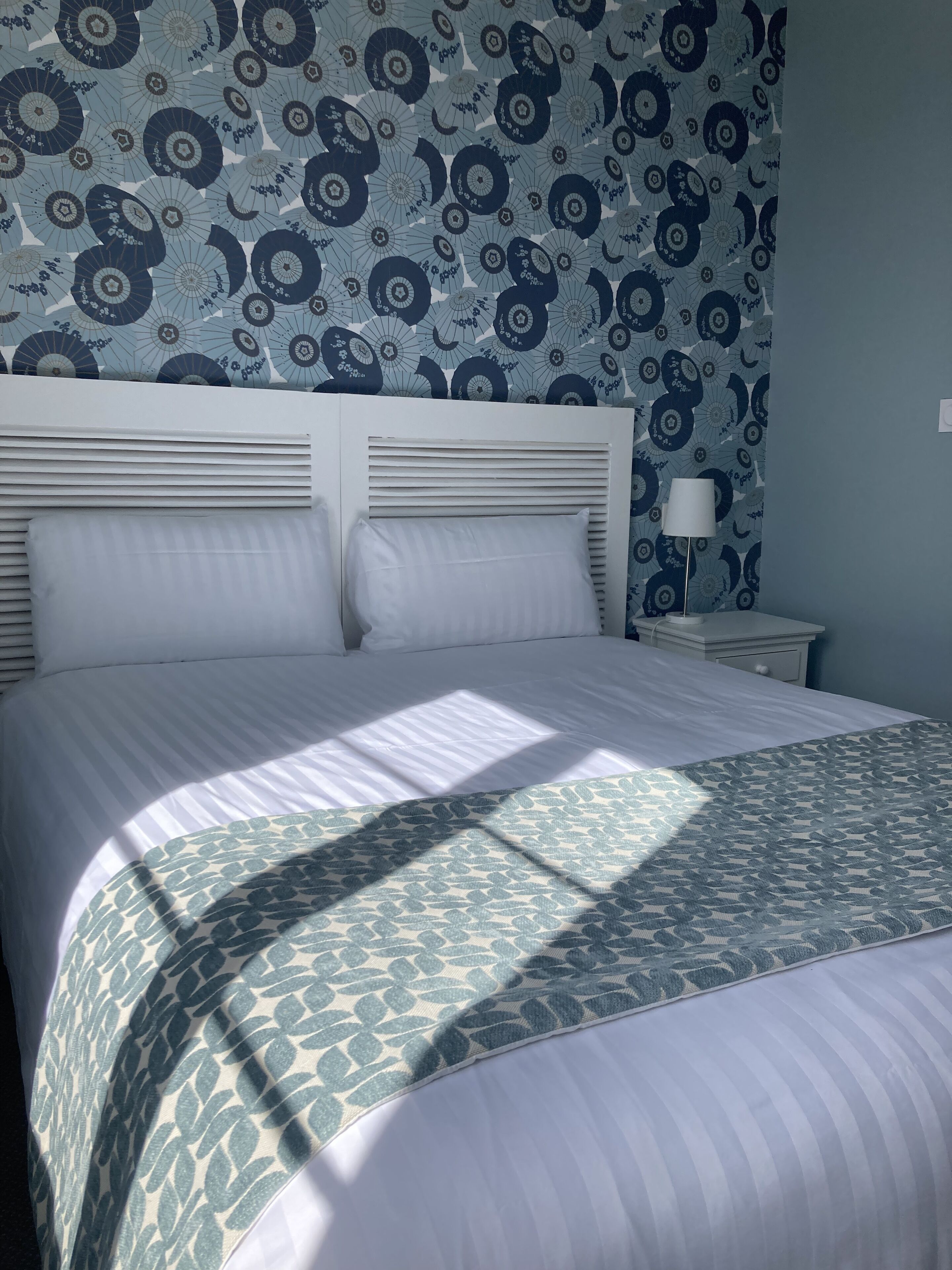 Un lit bien fait, recouvert d’une couvre-lit à motifs, avec une tête de lit blanche et un mur orné d’un papier peint à motifs floraux.