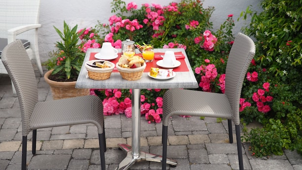 Une table mise pour le petit-déjeuner, avec des chaises en osier, une plante en pot et des fleurs roses en arrière-plan.