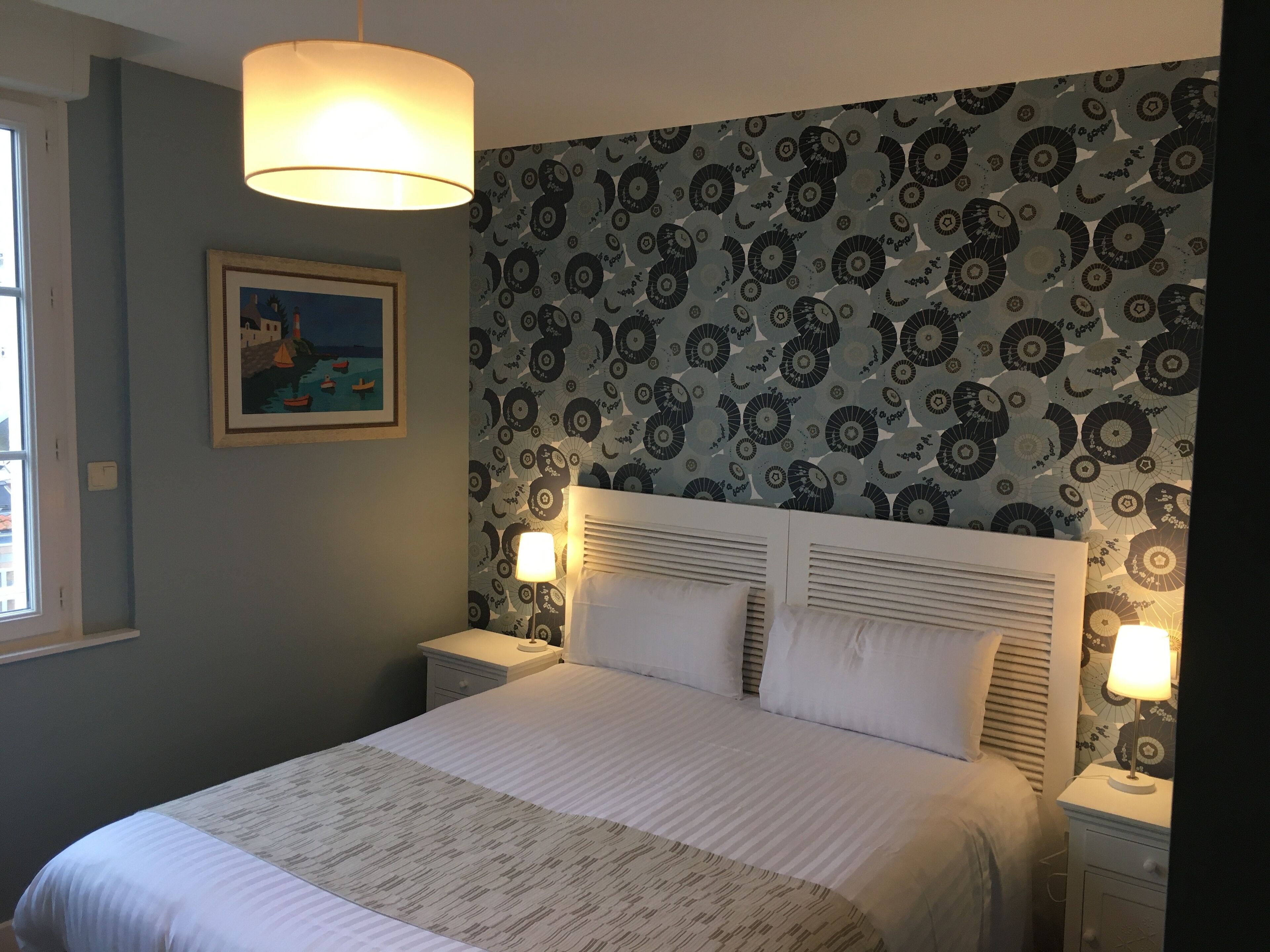 A neatly made bed with a patterned bedspread, two bedside lamps, a framed picture on the wall, and a patterned wallpaper.