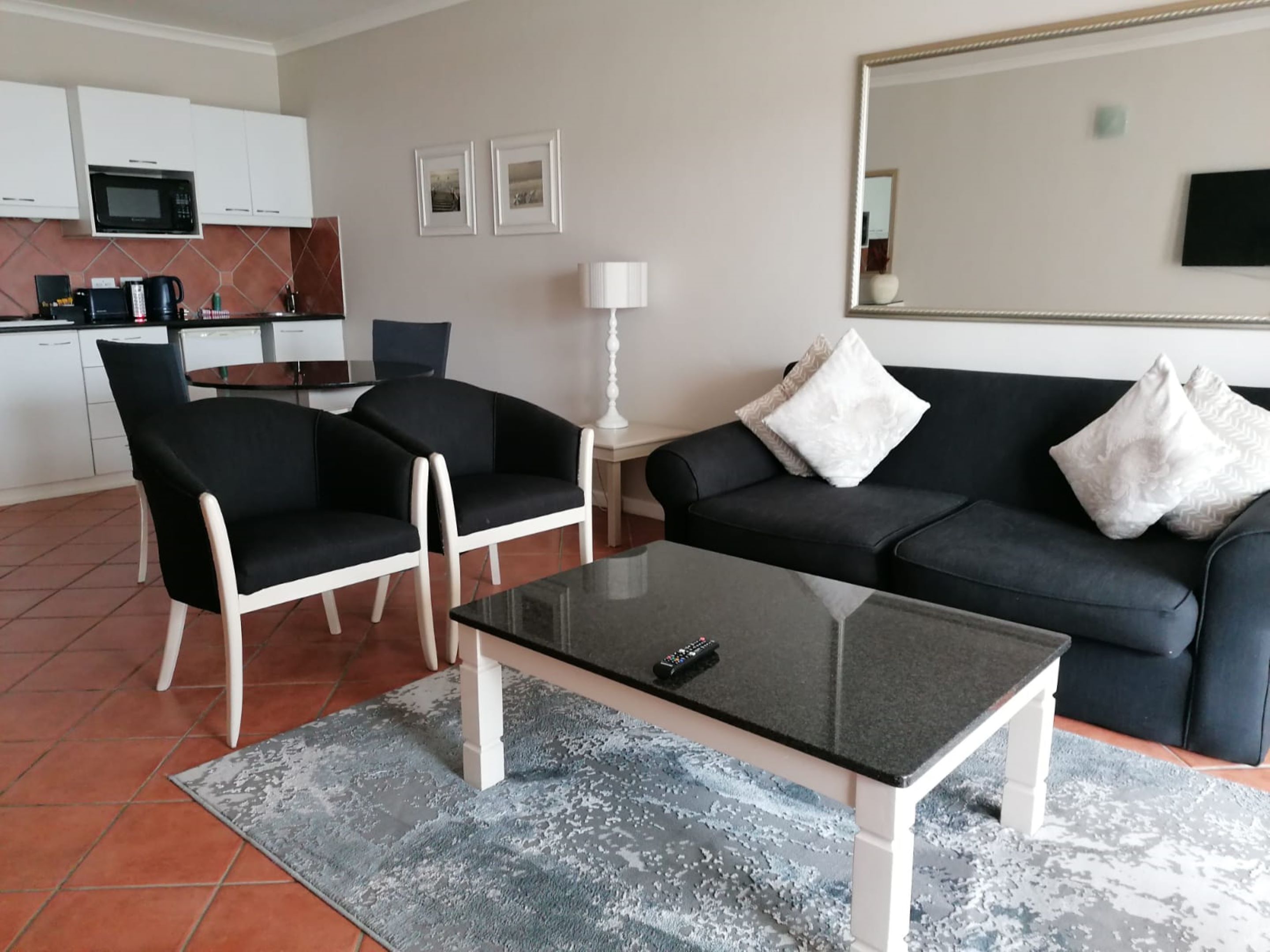 A modern living room with a black sofa, two black armchairs, a glass coffee table, and a kitchen area in the background.