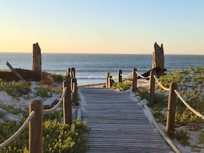 Un paseo marítimo de madera que conduce a la playa, rodeado de vegetación y postes de madera.