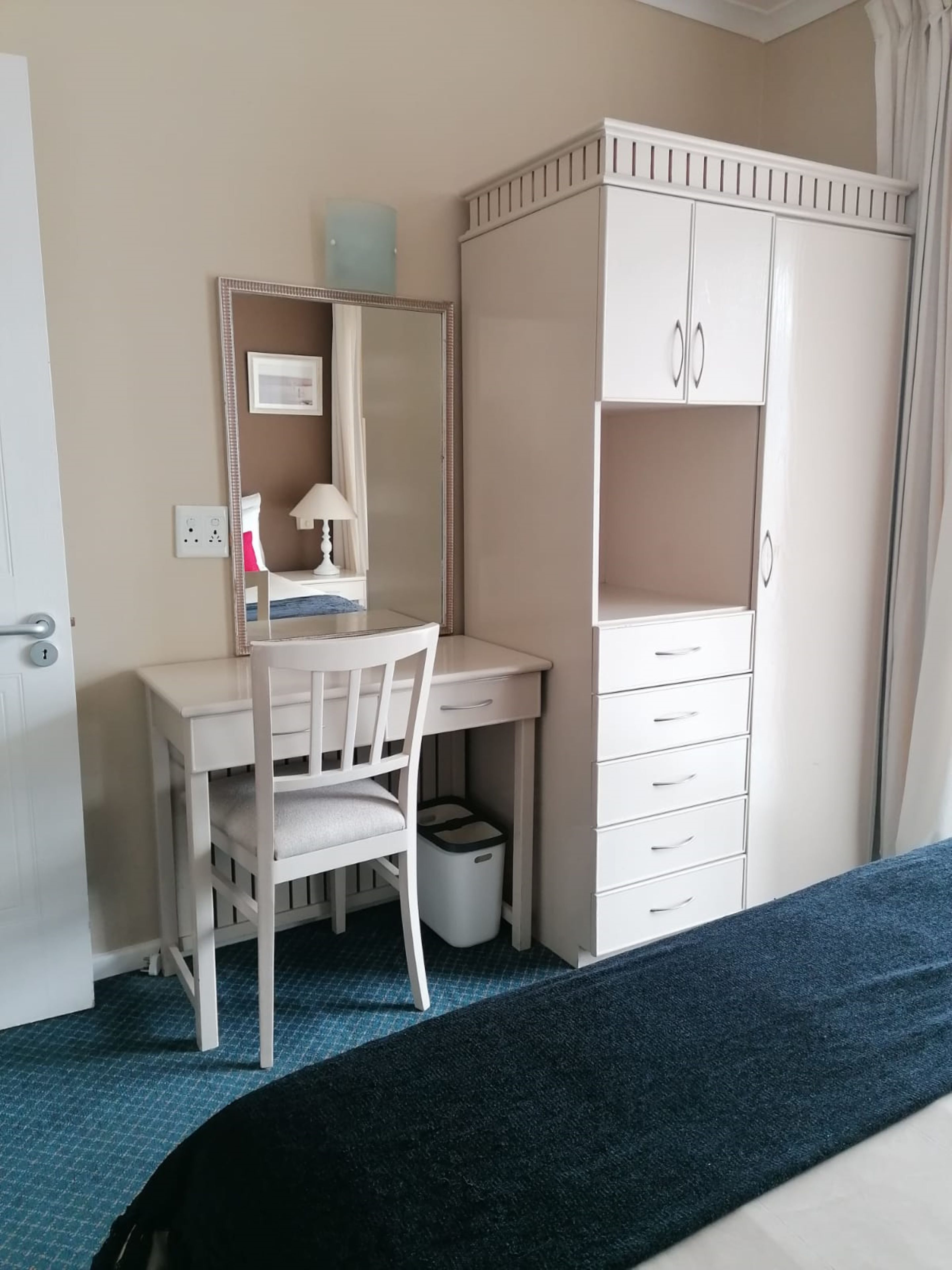A hotel room with a white desk, chair, and wardrobe. There is a mirror above the desk, a lamp on the desk, and a trash bin under the chair.