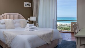 A hotel room with a bed, pillows, a bedside lamp, a view of the ocean, and a chair.