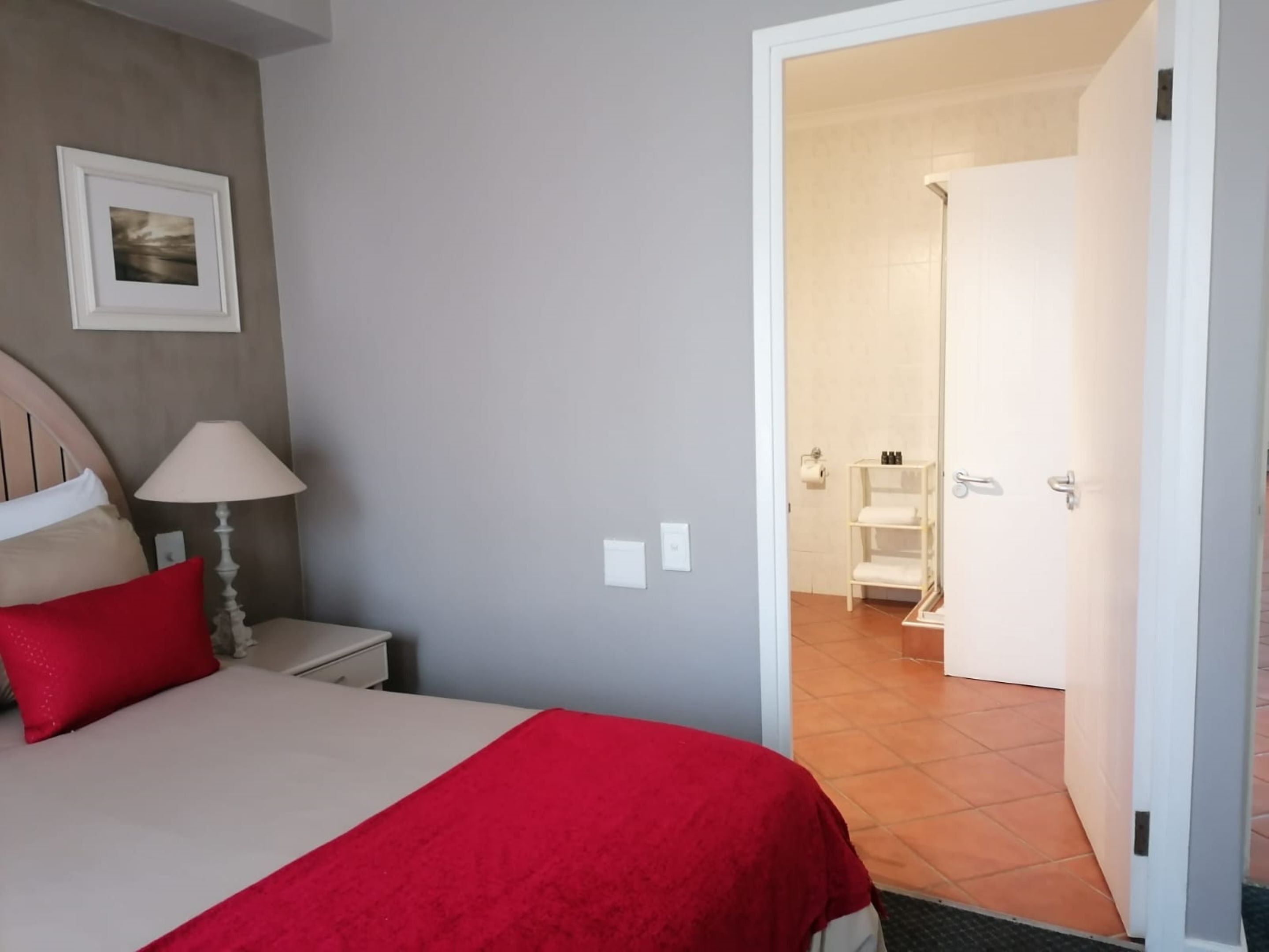 Un dormitorio con cama, mesita de noche, lámpara, almohada roja, un cuadro enmarcado y un baño visible a través de una puerta abierta.