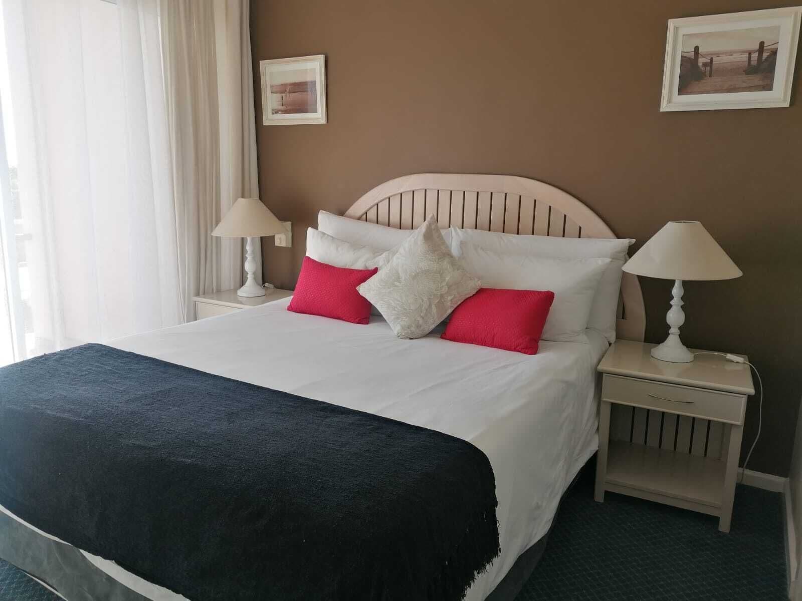 Un lit bien fait, agrémenté d’oreillers blancs et rouges, d’un plaid sombre et d’une tête de lit en bois. On y trouve deux tables de chevet avec des lampes et un petit tiroir.