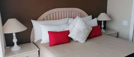 Una cama bien hecha con almohadas blancas y rojas, un cabecero de madera y dos lámparas de noche sobre mesitas blancas.