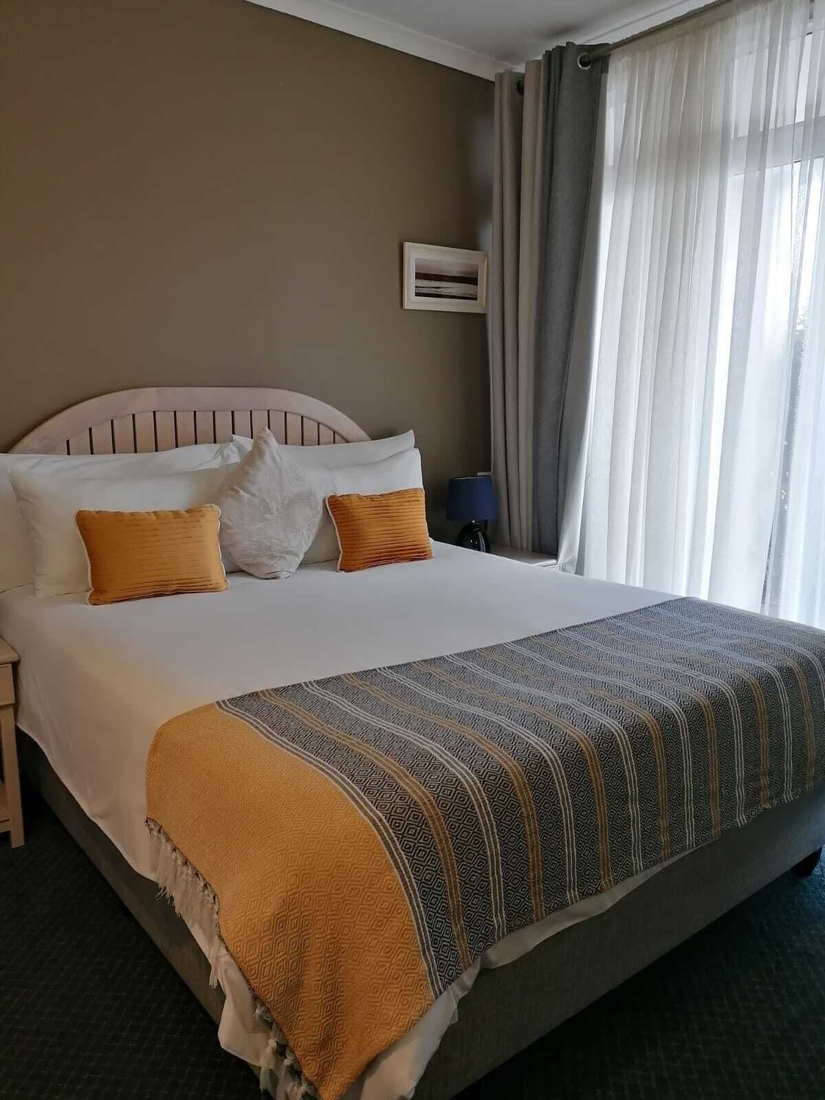 Una habitación de hotel con una cama grande, una mesita de noche, una lámpara y un ventanal con cortinas.