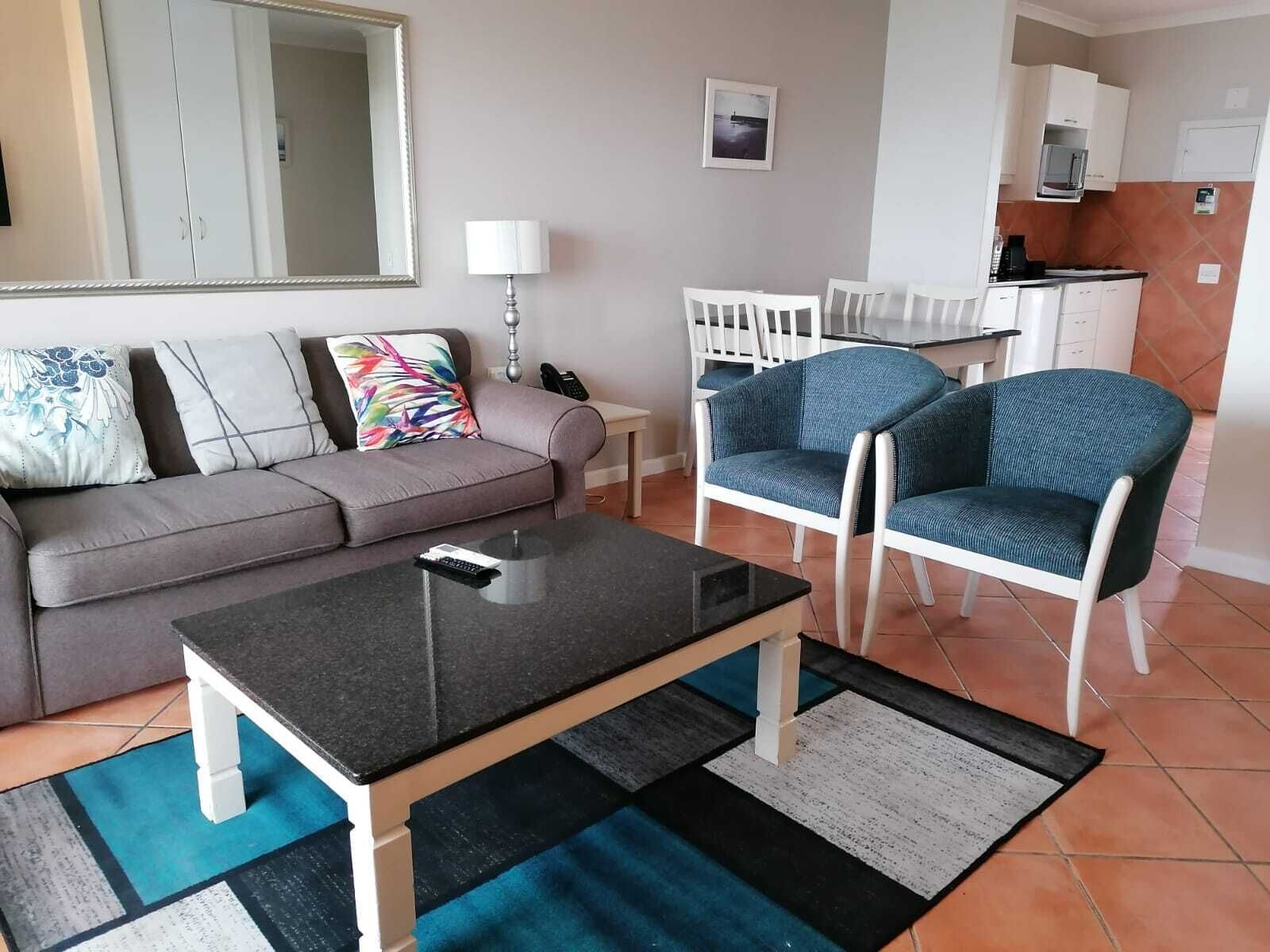 A living room with a grey sofa, a coffee table, and two blue armchairs. There is a kitchen area with white cabinets and a microwave.