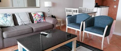 A living room with a grey sofa, a coffee table, and two blue armchairs. There is a kitchen area with white cabinets and a microwave.