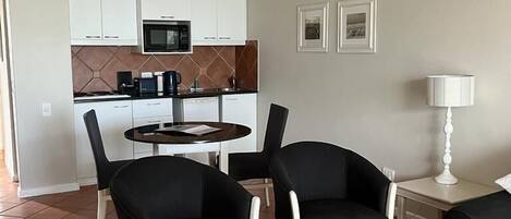 Una sala moderna con comedor, que incluye una cocina al fondo con armarios blancos y un microondas. Hay dos sillas negras y una mesa redonda pequeña.