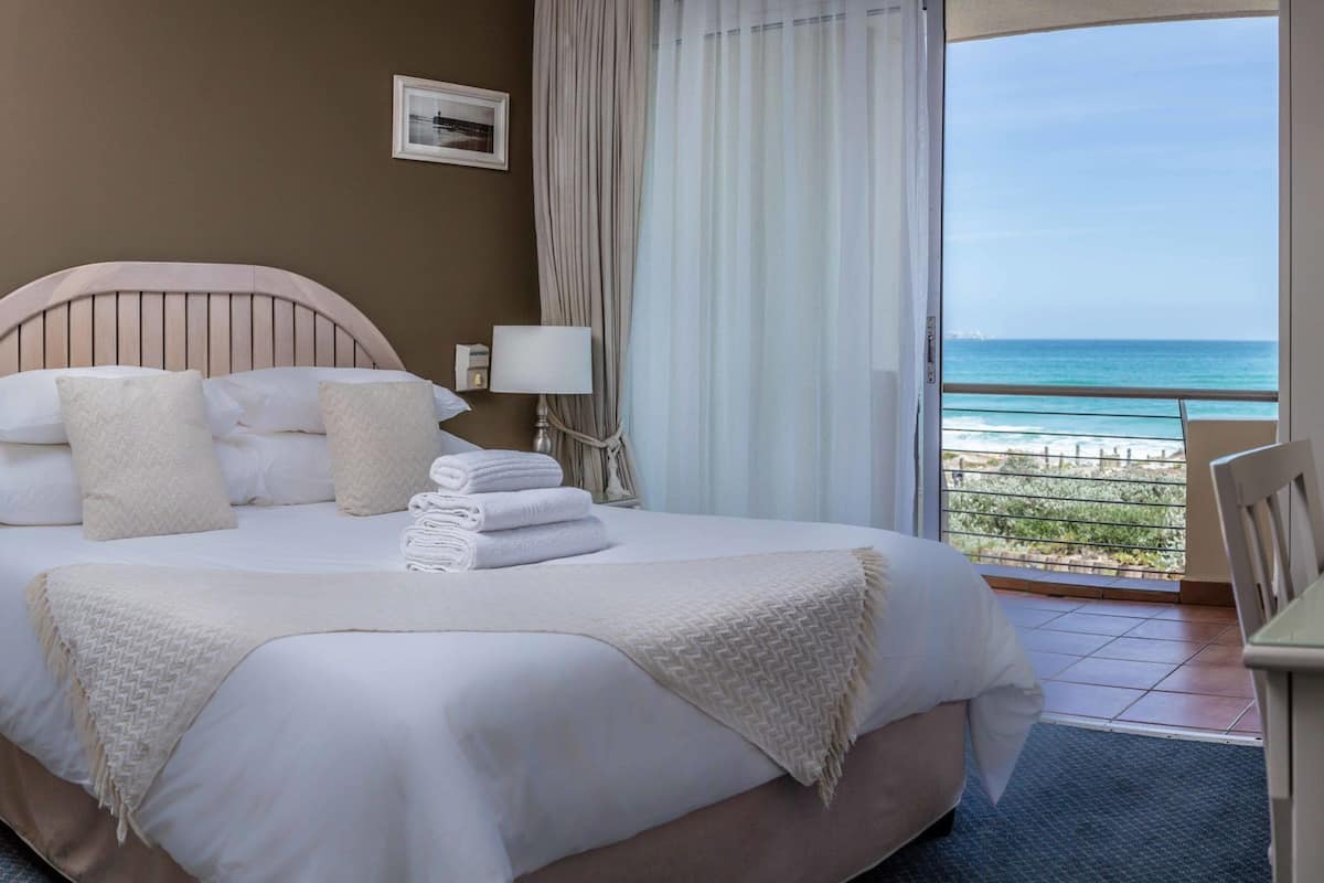 Een hotelkamer met een bed, nachtkastje, lamp, uitzicht op de oceaan en een balkon.