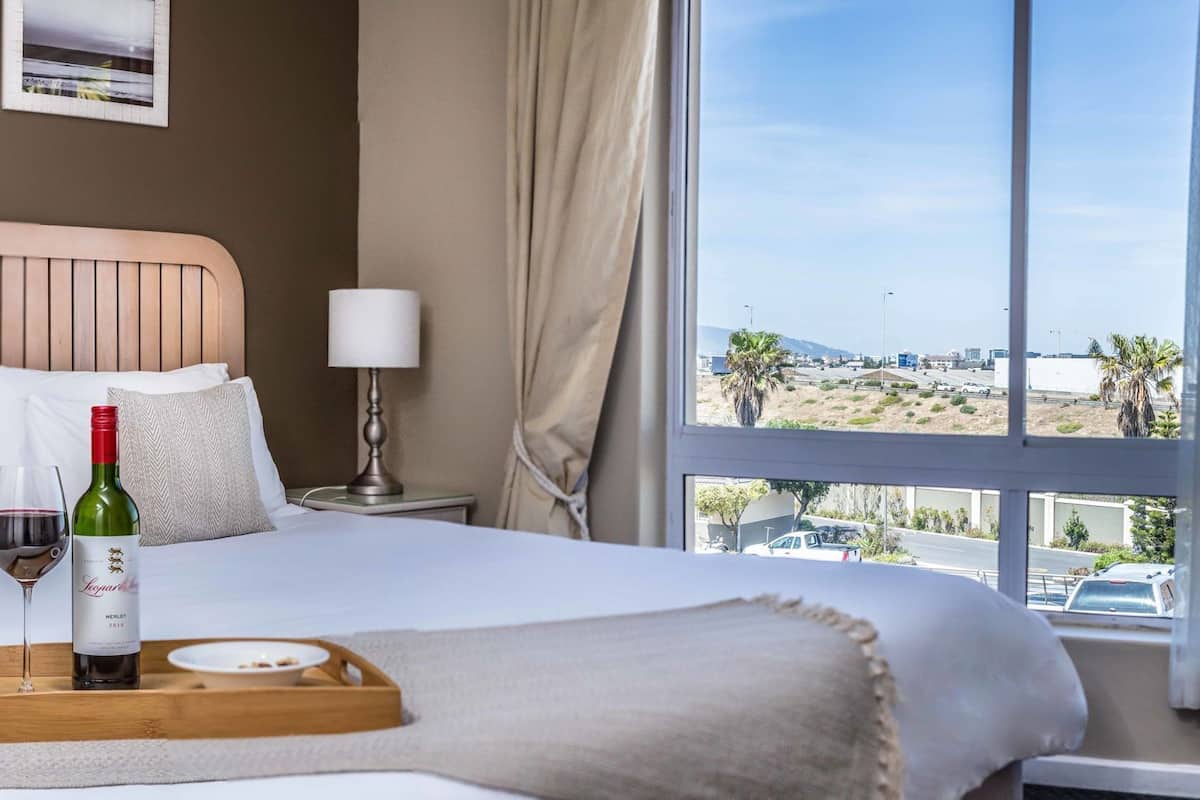 Een hotelkamer met een bed, een fles wijn, een glas wijn, een dienblad en uitzicht op strand en stad.