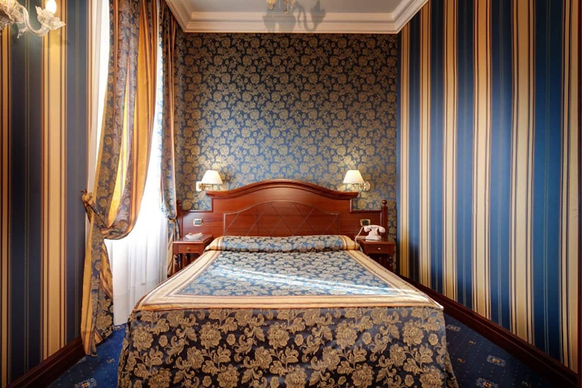 A bedroom with a large bed, two bedside tables with lamps, a wooden headboard, and a patterned wallpaper.