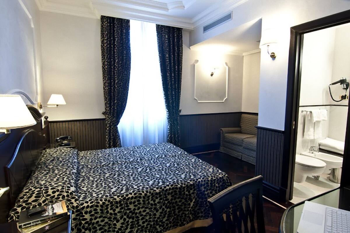 A hotel room with a bed, a desk with a lamp, a chair, a window with curtains, a bathroom visible through a glass door, and a wall-mounted light fixture.