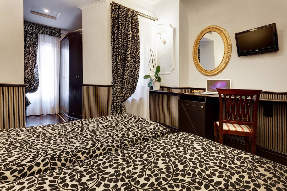 A hotel room with a patterned bedspread, a wooden chair, a television, a mirror, and a potted plant.