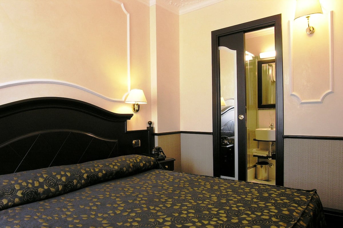 A hotel room with a bed, a nightstand, a lamp, a mirror, and a door leading to a bathroom.
