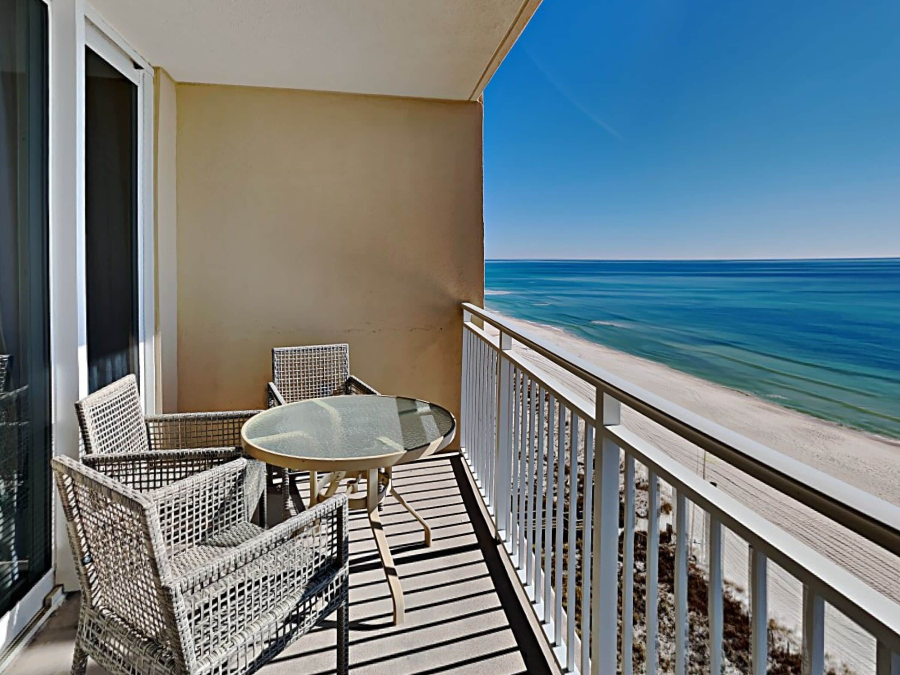 Een balkon met rieten meubilair en een glazen tafel met uitzicht op een strand.