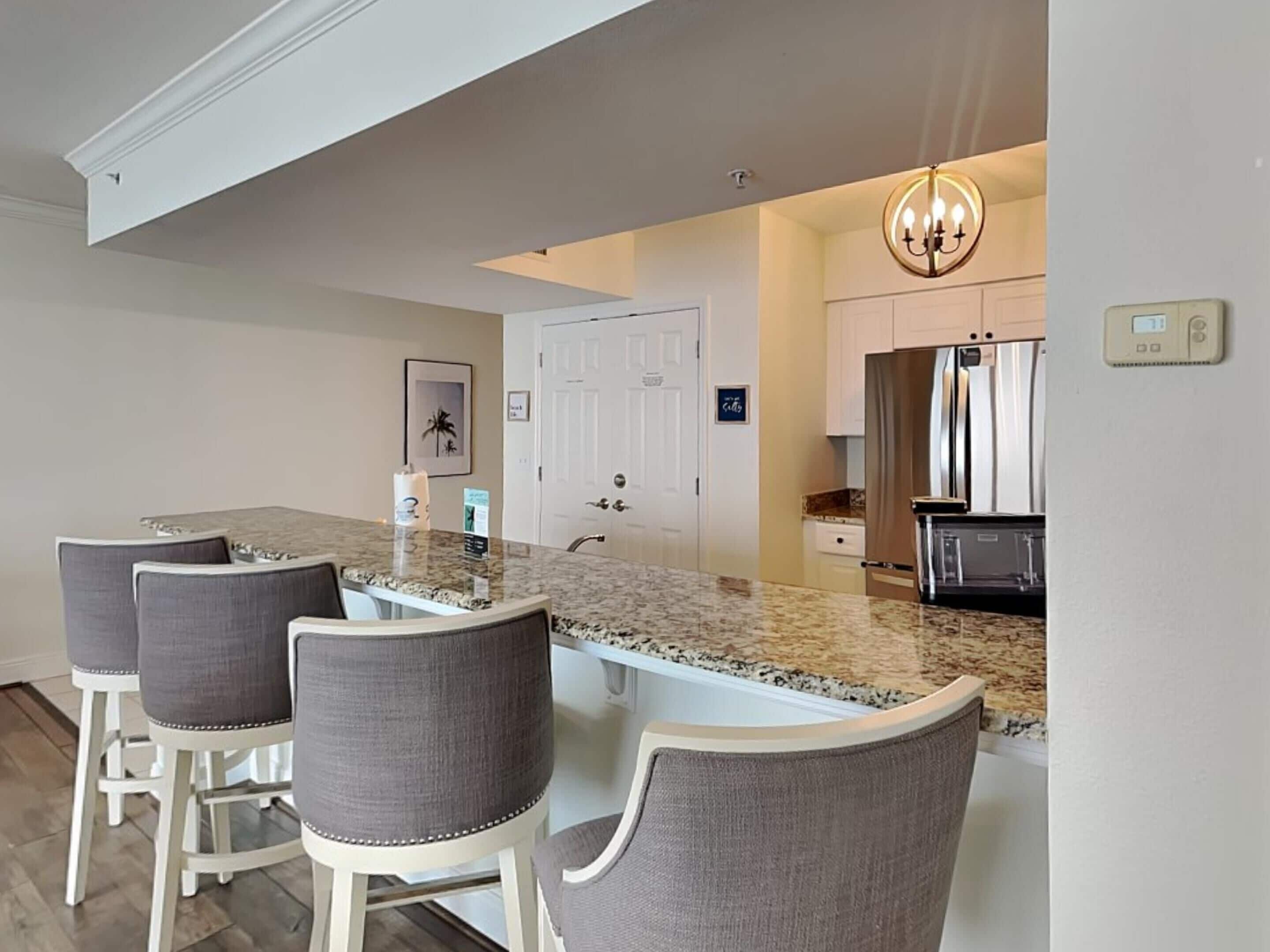 A modern kitchen with a granite countertop, white cabinets, and a stainless steel refrigerator. There are three bar stools with gray fabric seats.