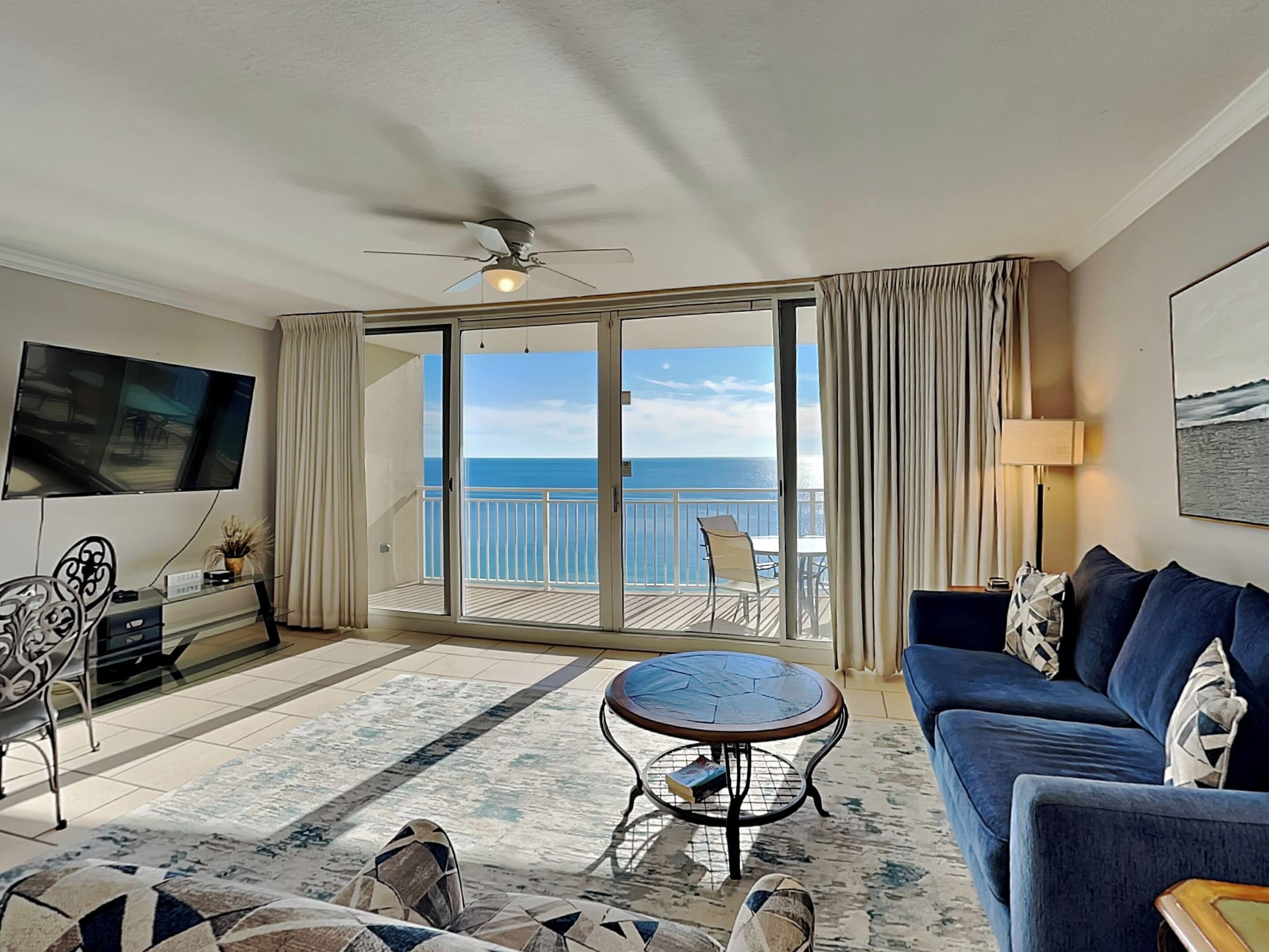 A living room with a sofa, coffee table, and TV. It has a balcony with a view of the ocean.