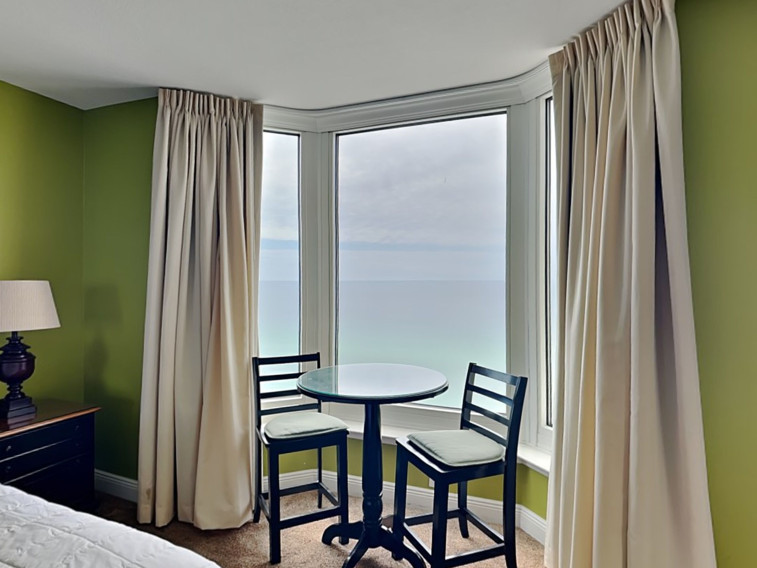 A hotel room with a corner table and two chairs, a lamp, and a view of the ocean through large windows.