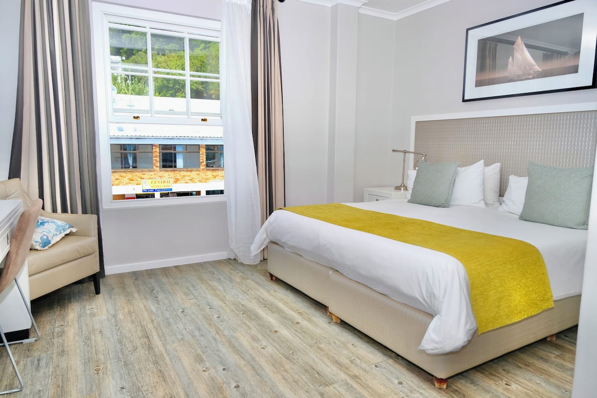 A hotel room with a bed, a chair, a window with curtains, a framed picture on the wall, and a wooden floor.