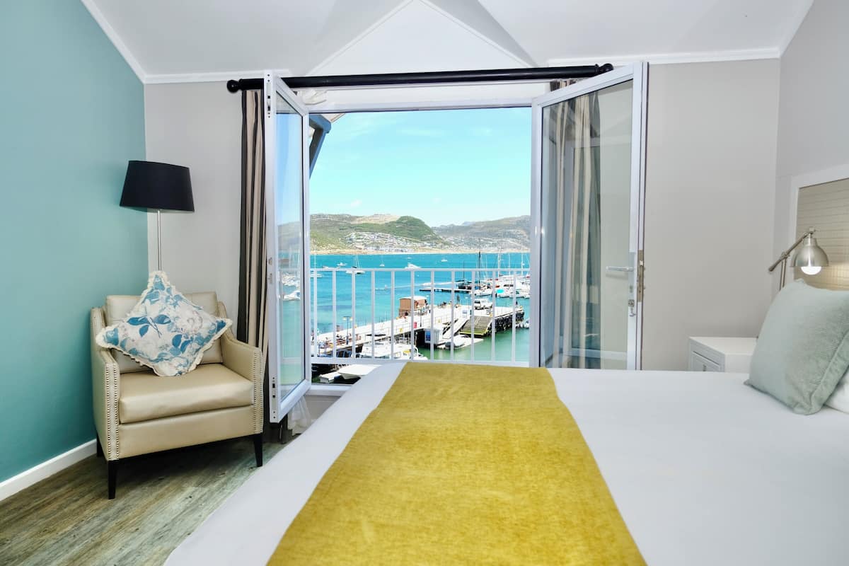 A bedroom with a bed, a chair, a lamp, and a view of the sea and boats through a glass door.