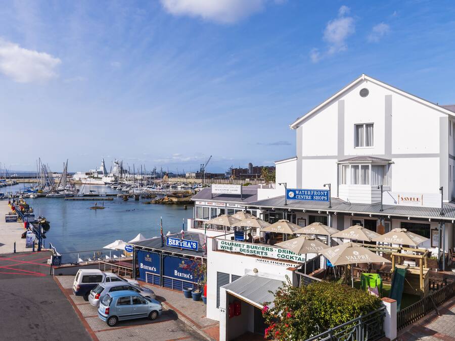 Une propriété située au bord de l’eau, avec un bâtiment blanc, une marina remplie de bateaux et un espace extérieur aménagé avec des parasols.