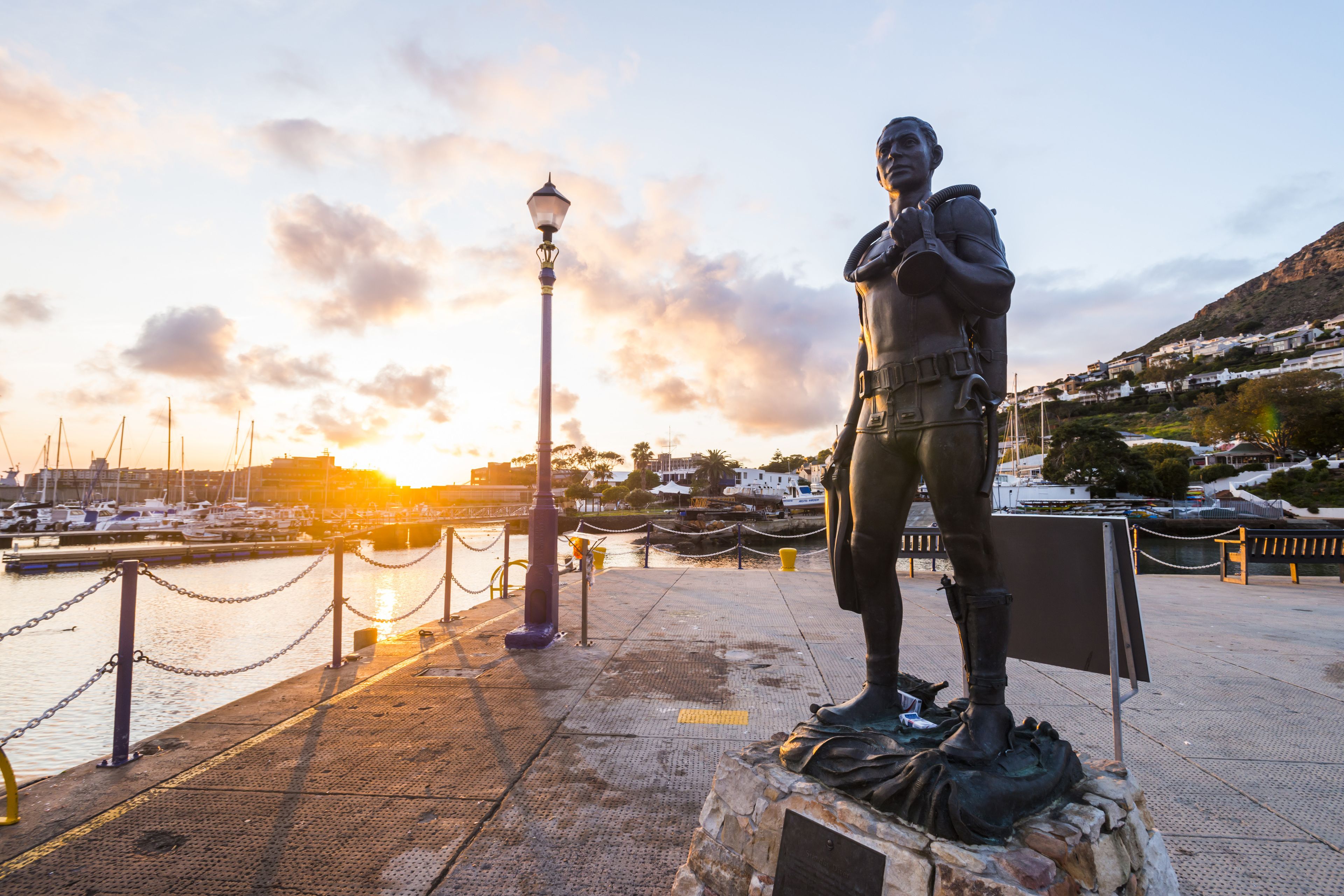 A statue of a person in uniform standing on a pedestal by a waterfront with a sunset in the background.