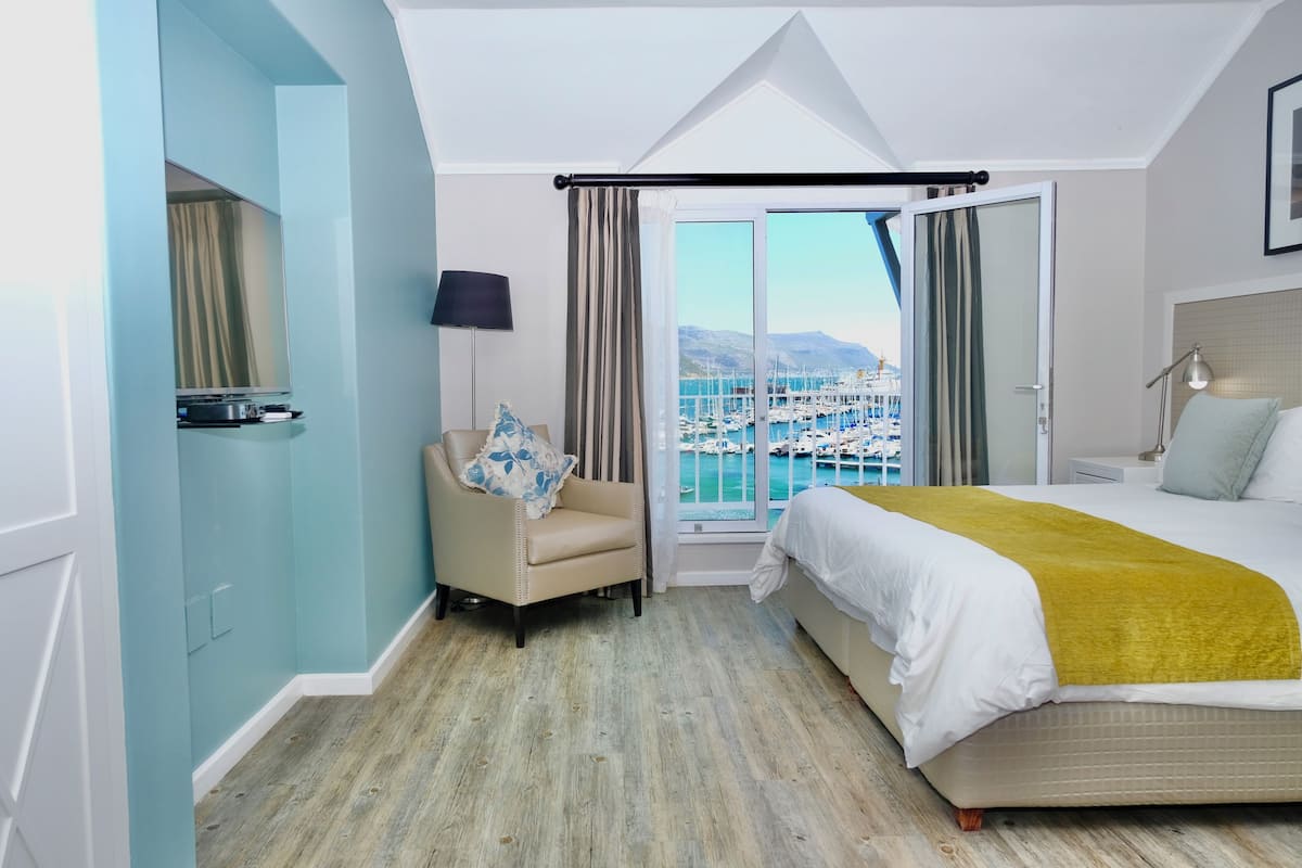 A hotel room with a bed, a chair, a desk, a lamp, and a view of a marina.