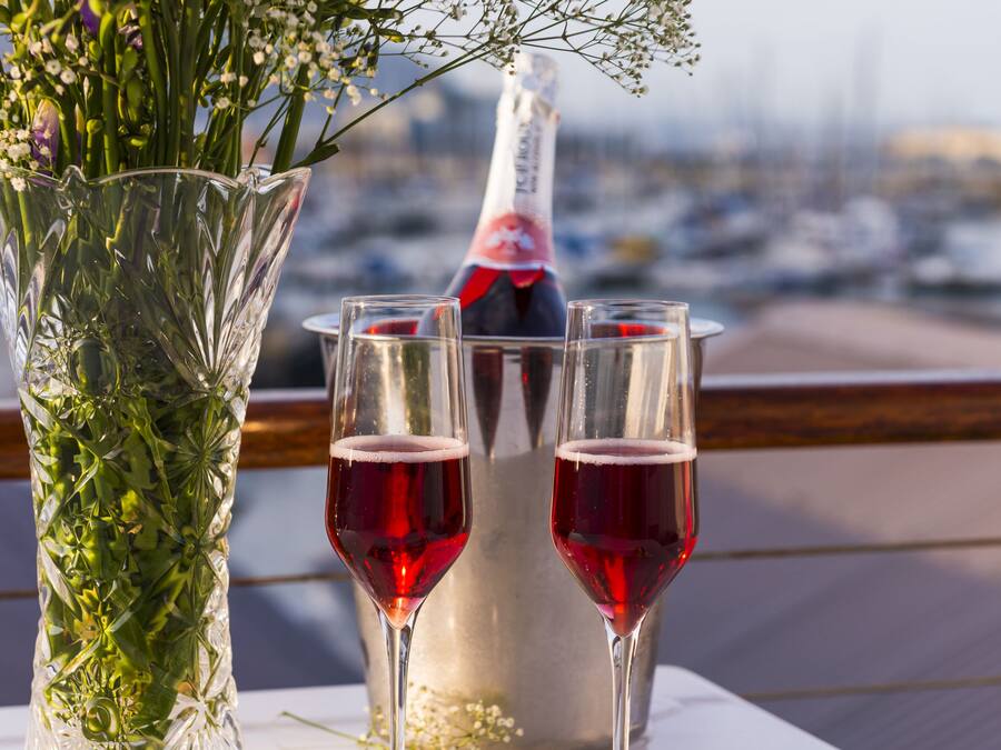 Una mesa con un florero de vidrio con flores moradas, dos copas de vino tinto, una botella de champán y una selección de bombones y fresas.