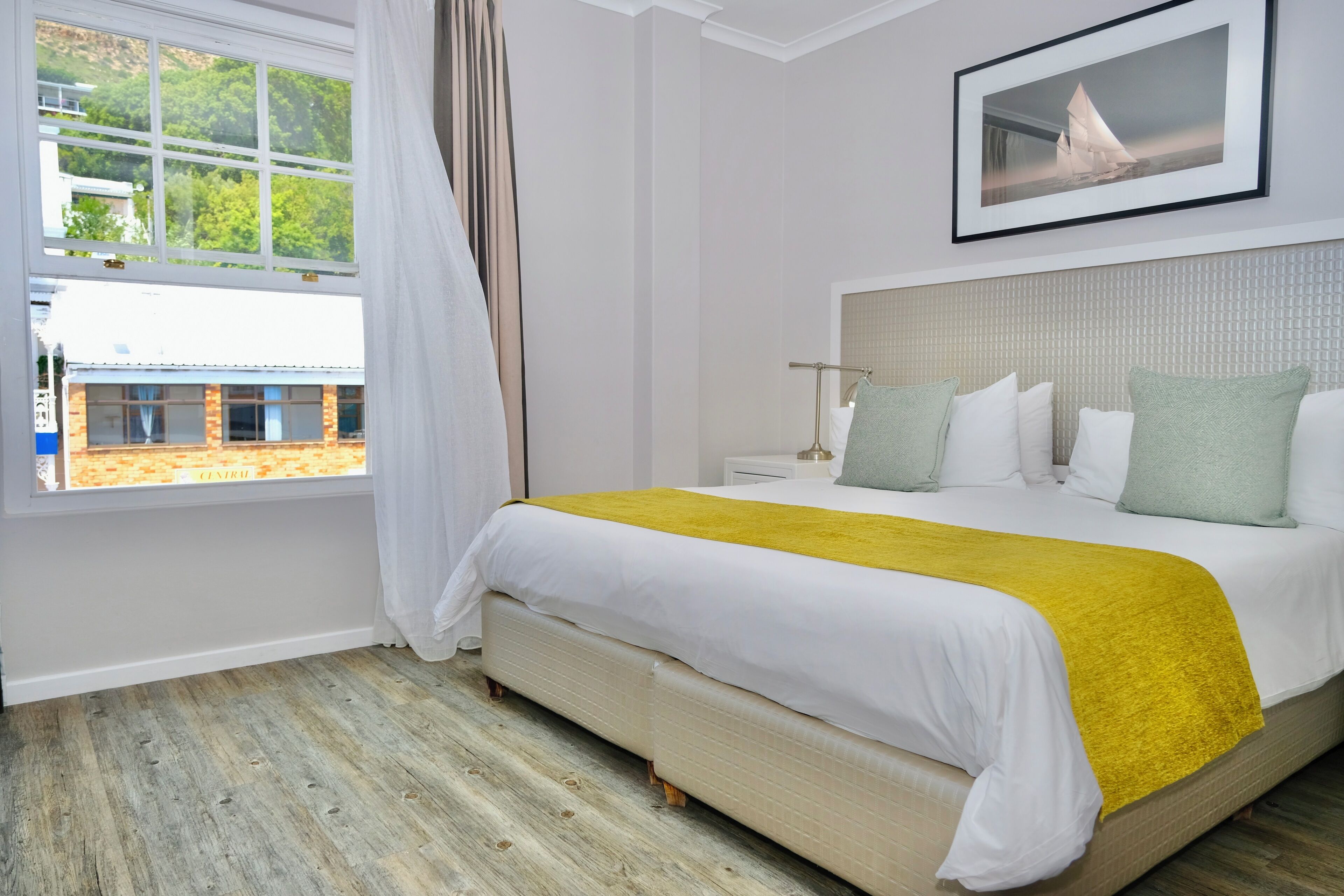 Habitación de hotel con una cama grande, una ventana con cortinas, una mesita de noche y un cuadro enmarcado en la pared.