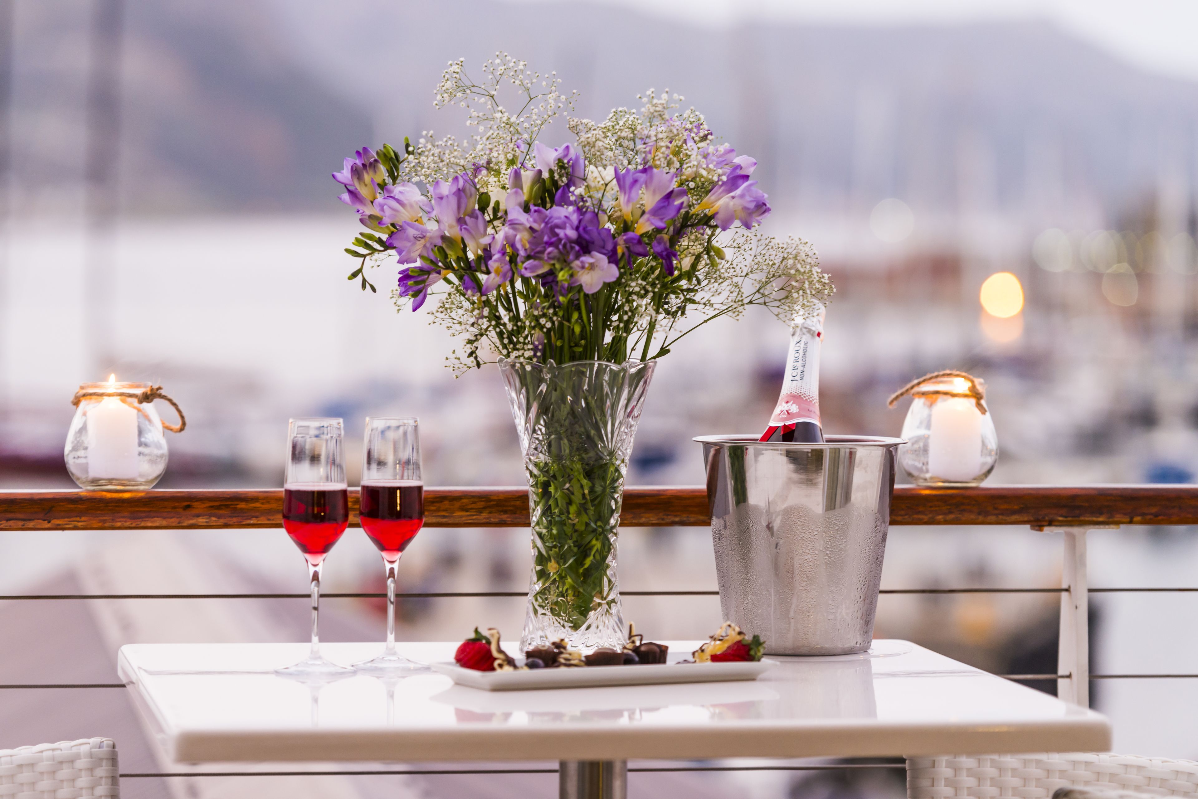 A table with a vase of purple flowers, two glasses of red wine, and a silver bucket with a candle.