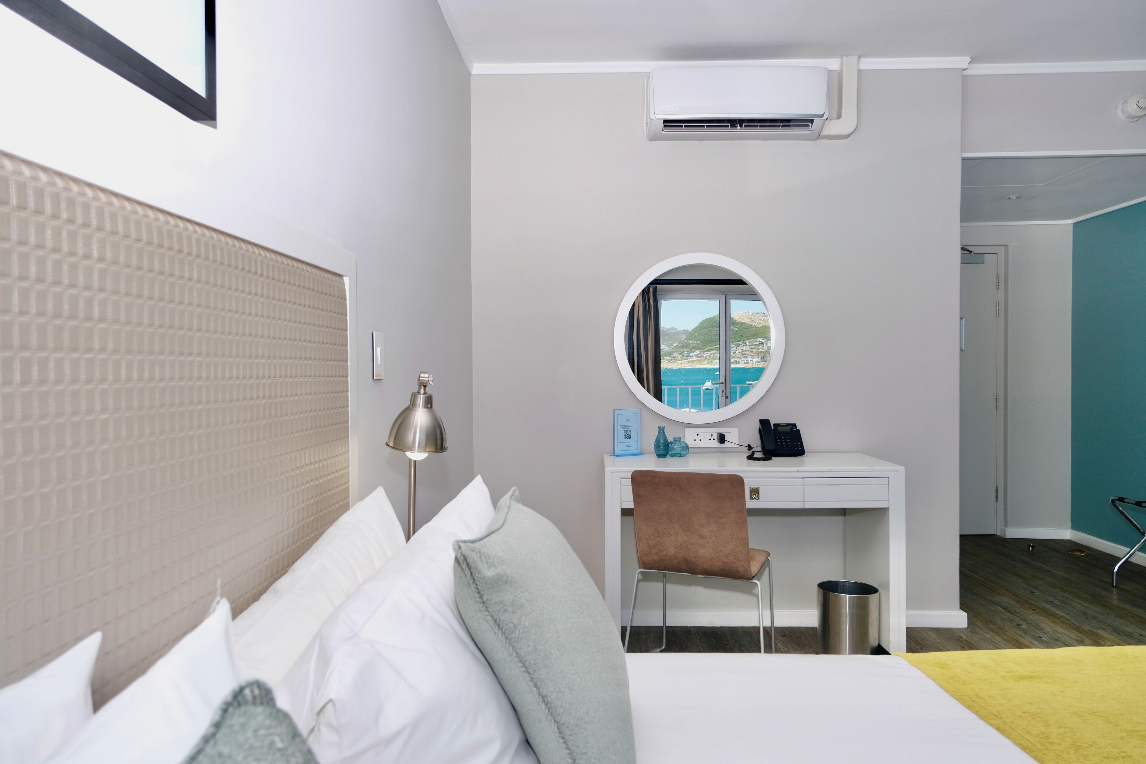 Une chambre d’hôtel avec un lit, un bureau et une chaise, un miroir rond et une vue sur la mer.
