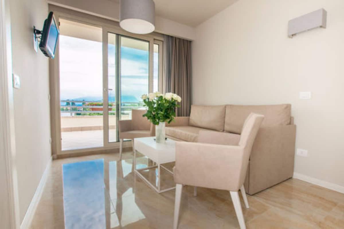 A modern living room with a beige sofa, a small table with a vase of flowers, and a view of the outdoors through a sliding glass door.