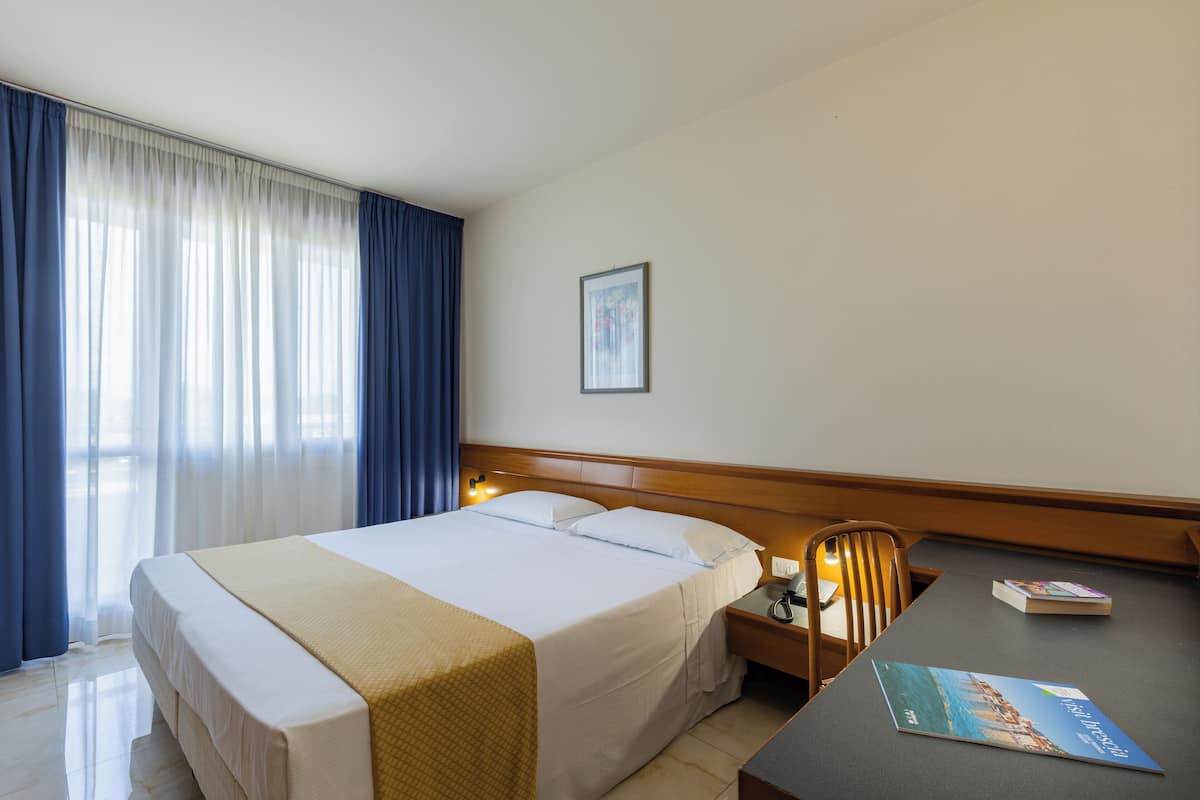 A hotel room with a bed, a desk, a chair, a framed picture on the wall, and a window with curtains.