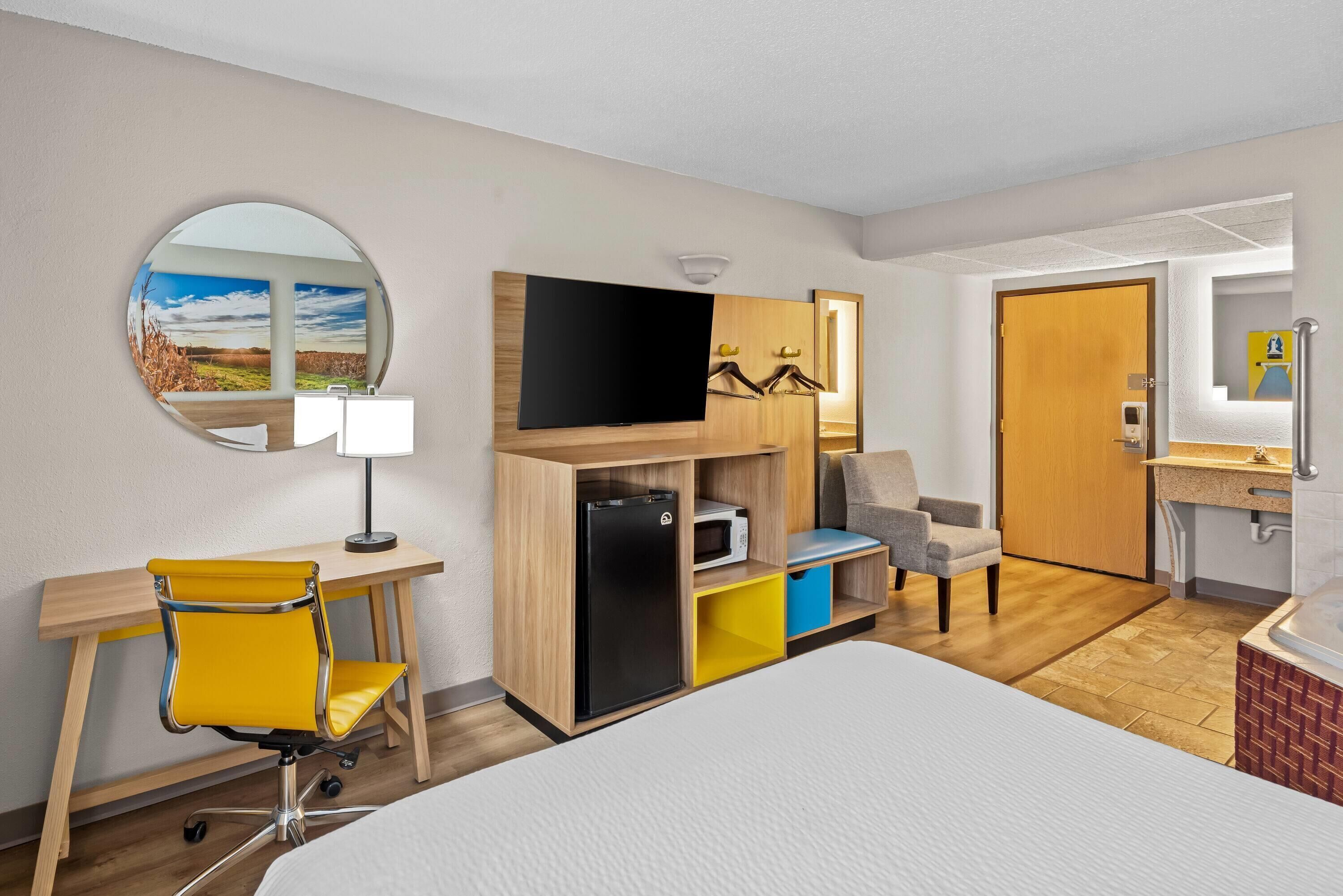 Habitación de hotel con cama, escritorio con silla, televisor, microondas, refrigerador pequeño, espejo y baño con lavamanos e inodoro.