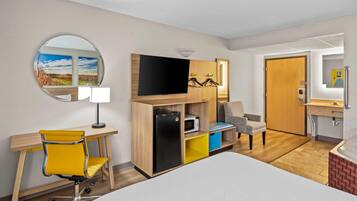 Habitación de hotel con cama, escritorio con silla, televisor, microondas, refrigerador pequeño, espejo y baño con lavamanos e inodoro.