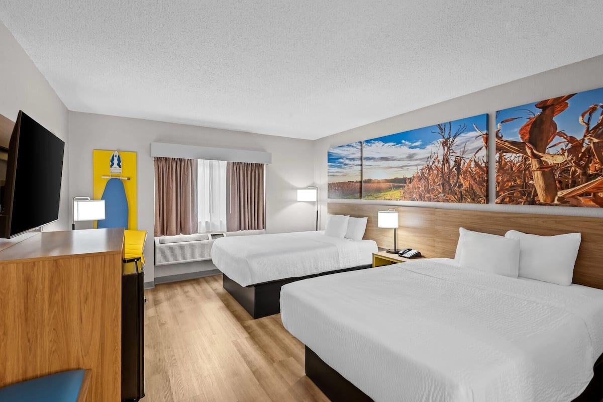 A hotel room with two beds, a desk, a TV, and a large painting of a field.