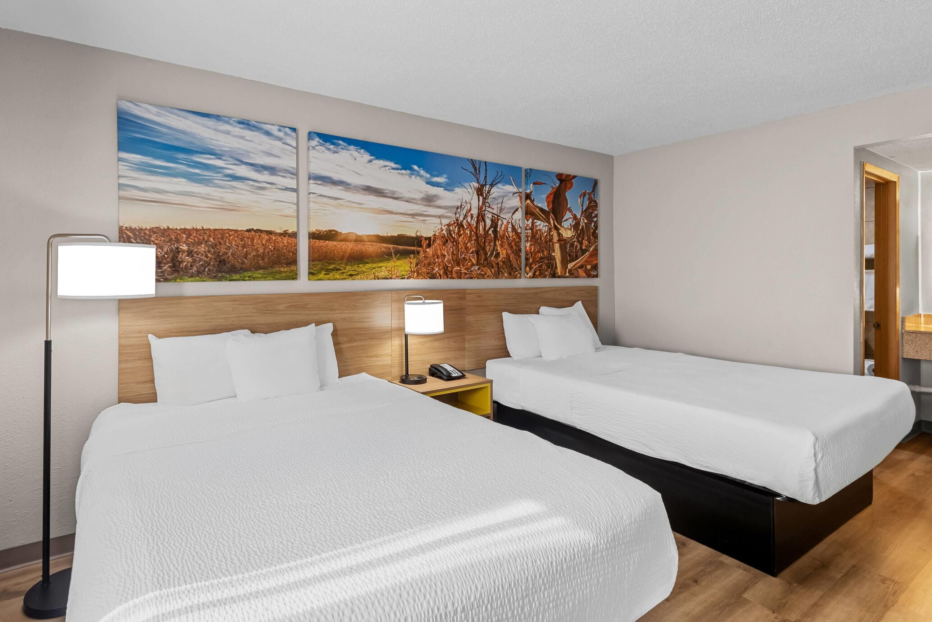 Habitación de hotel con dos camas, cabecera de madera, mesita de noche con lámpara y un cuadro grande de un paisaje en la pared.